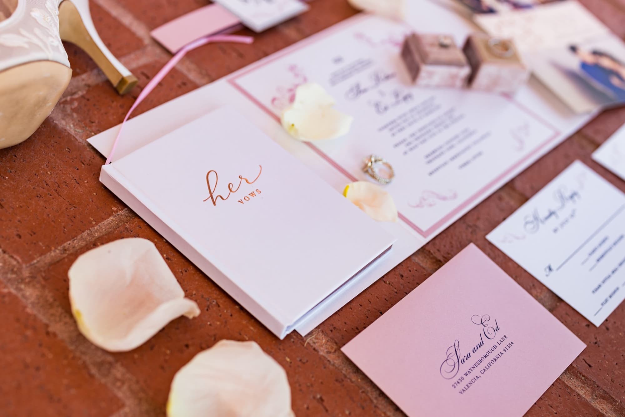 Flat lay of a blush wedding invitation suite addressed to Sara and Ed with a her vows book, lace bridal heel, and engagement ring on brick at Sherwood Country Club
