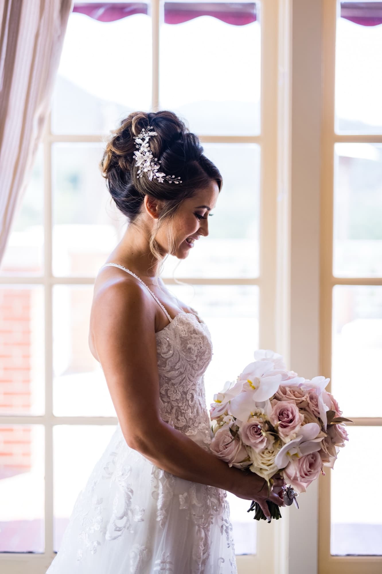 Bride in a lace A-line gown holding a blush rose and orchid bouquet in the window light of the Sherwood Country Club bridal suite