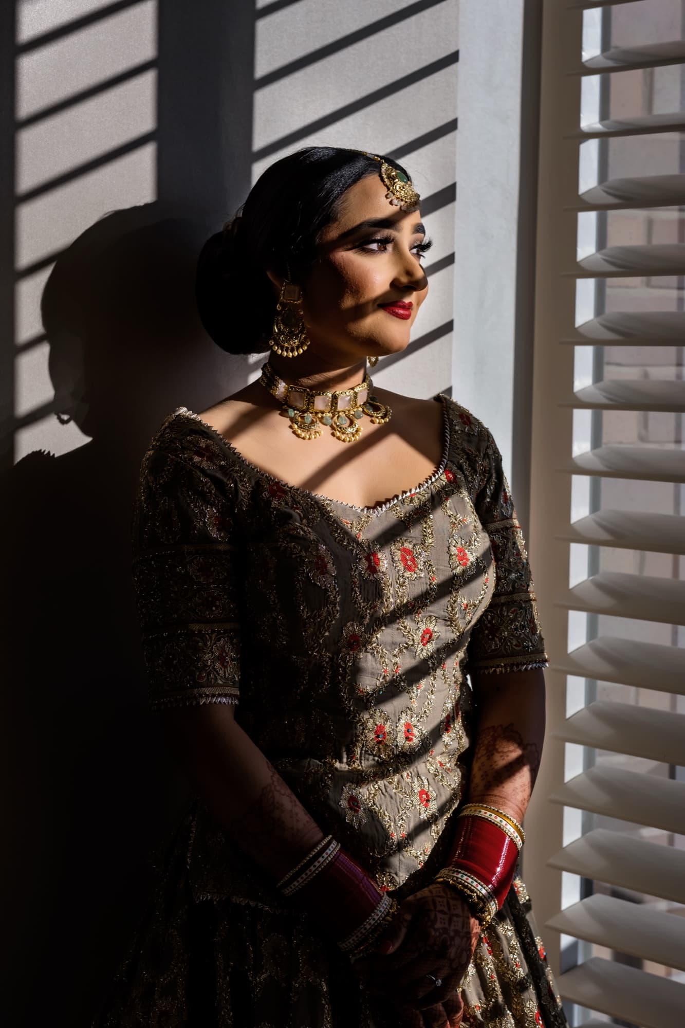 Harmen in her embroidered lehenga and gold jewelry lit by slatted window light on the wedding morning