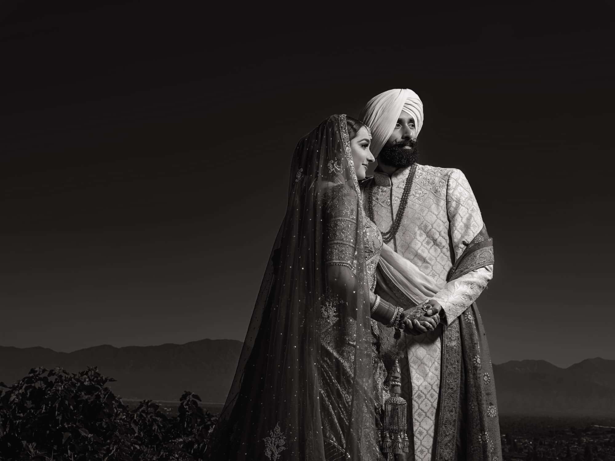 Dusk black and white portrait of Harmen and Amaritpal beneath a wide darkening sky, her embroidered dupatta trailing