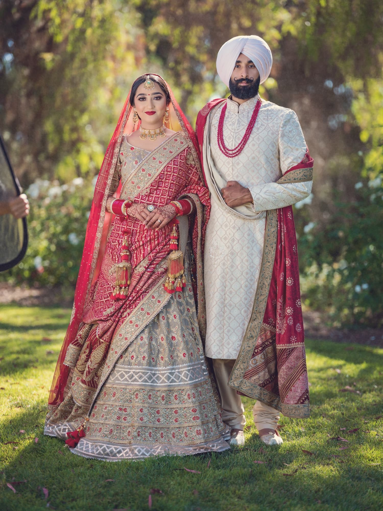 Harmen in an intricately embroidered red lehenga and Amaritpal in an ivory sherwani with a red dupatta standing together in a rose garden, photographed by Michael Anthony Photography