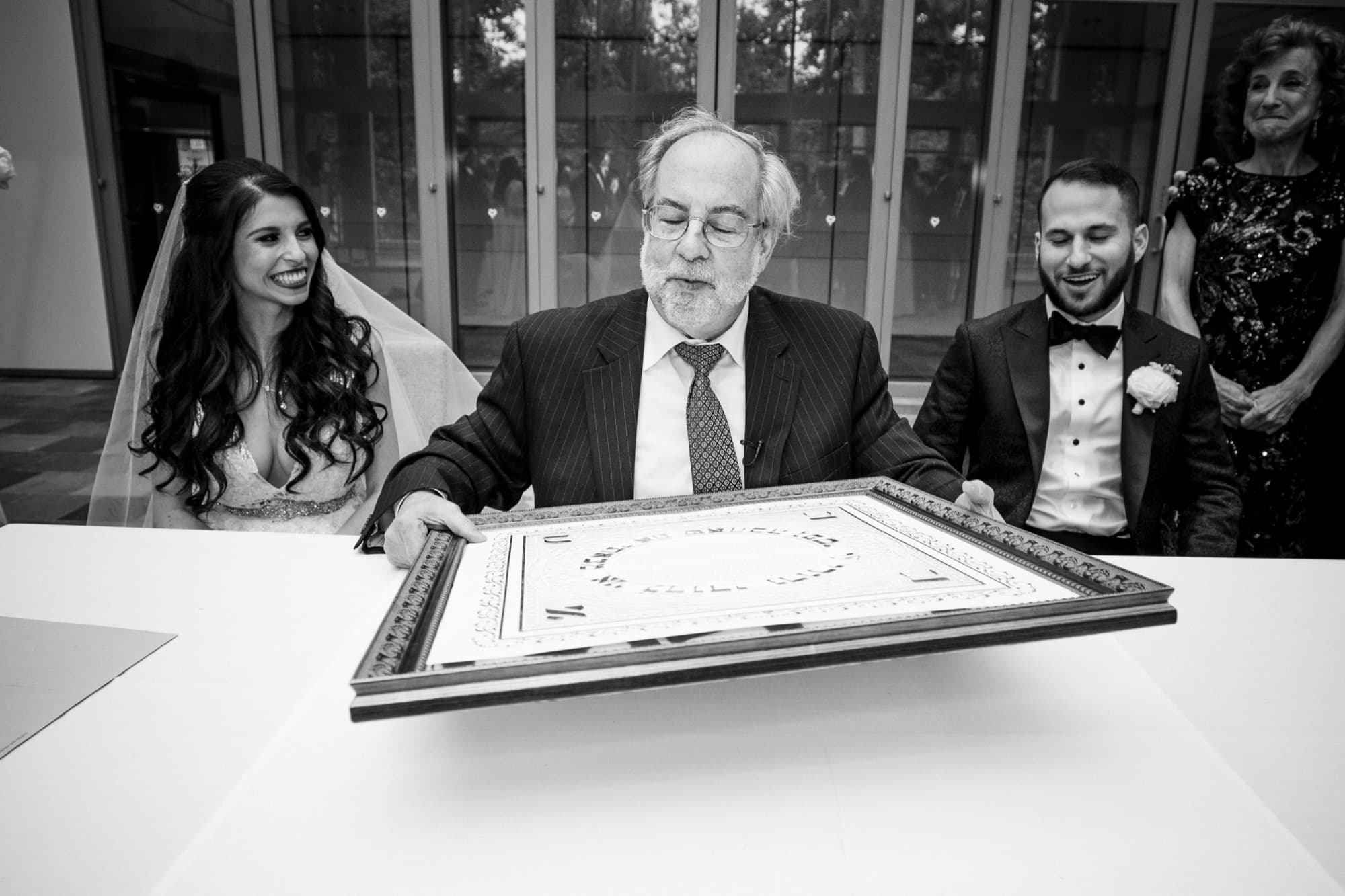 Black and white photograph of the rabbi presenting the ketubah as Rachel and Jeff look on with family
