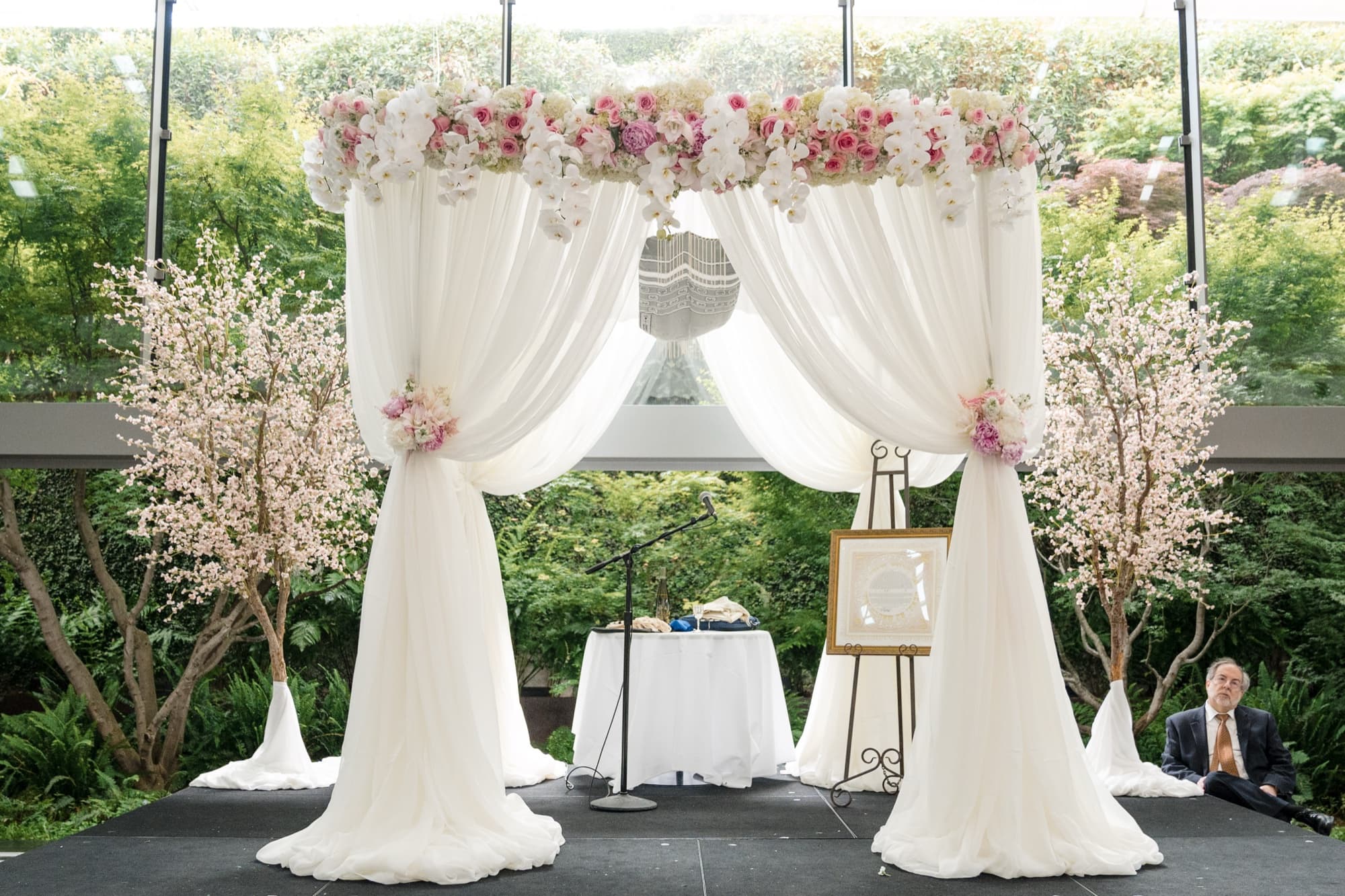 The flower-crowned chuppah in the Skirball's glass pavilion, white drapery and blossom branches with gardens beyond