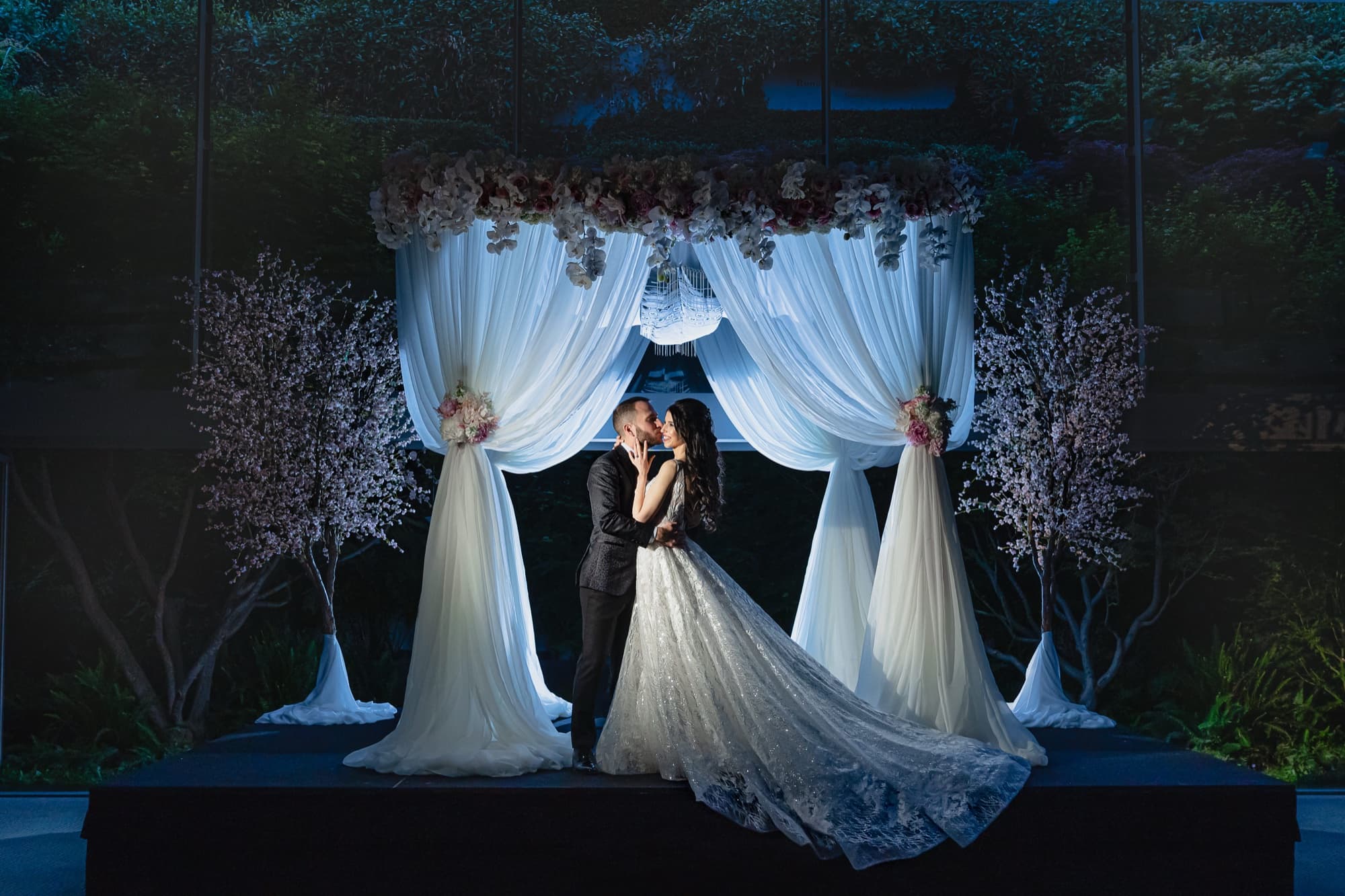 Rachel and Jeff kissing beneath their illuminated flower-crowned chuppah at night at the Skirball Cultural Center, her lace train spread across the platform, photographed by Michael Anthony Photography