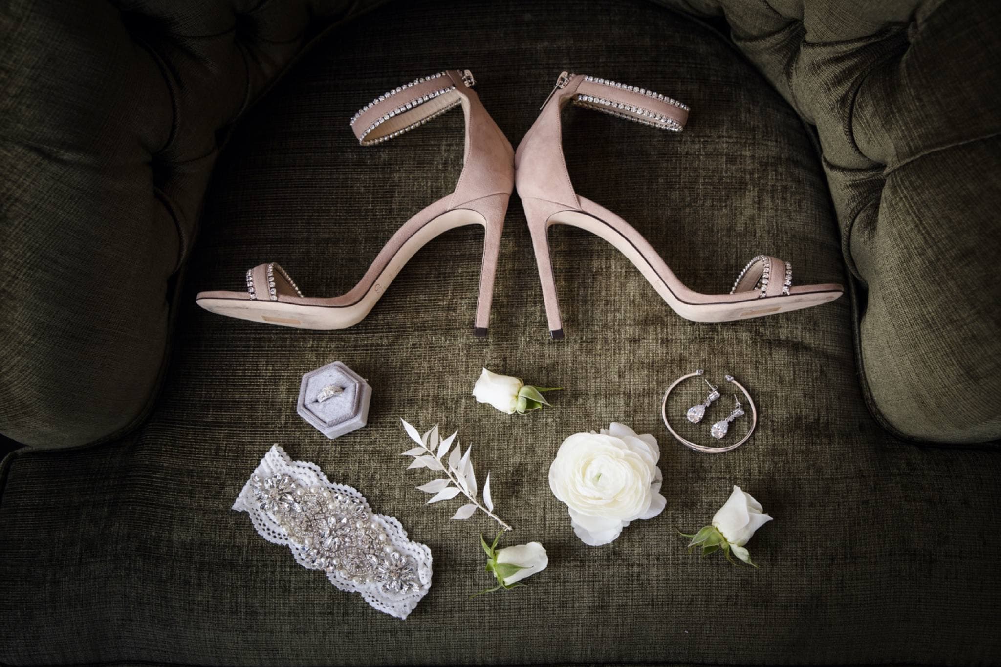 Blush heels, a crystal garter, rings, and white roses arranged on green velvet before the wedding