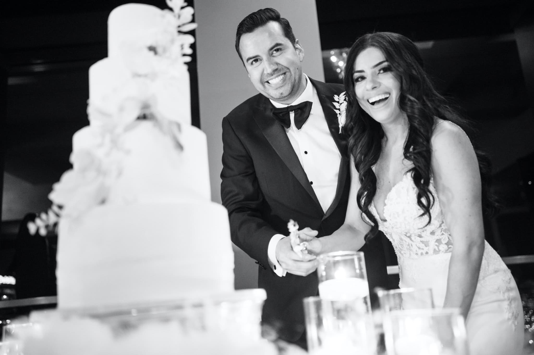 Black and white photograph of Christina and Ryan laughing together as they cut their tiered wedding cake