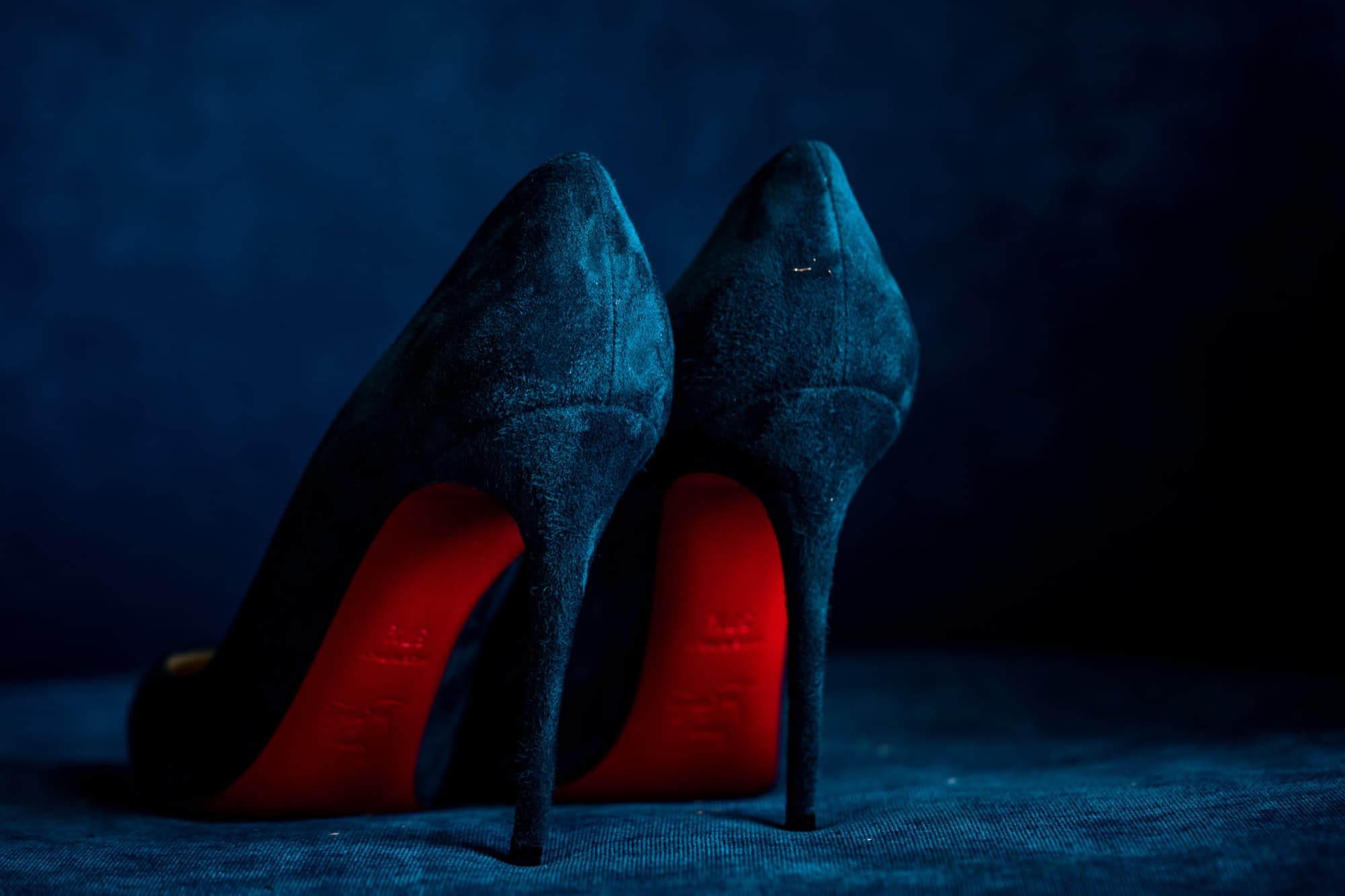Midnight blue suede heels photographed sole-first showing their red bottoms against a blue backdrop