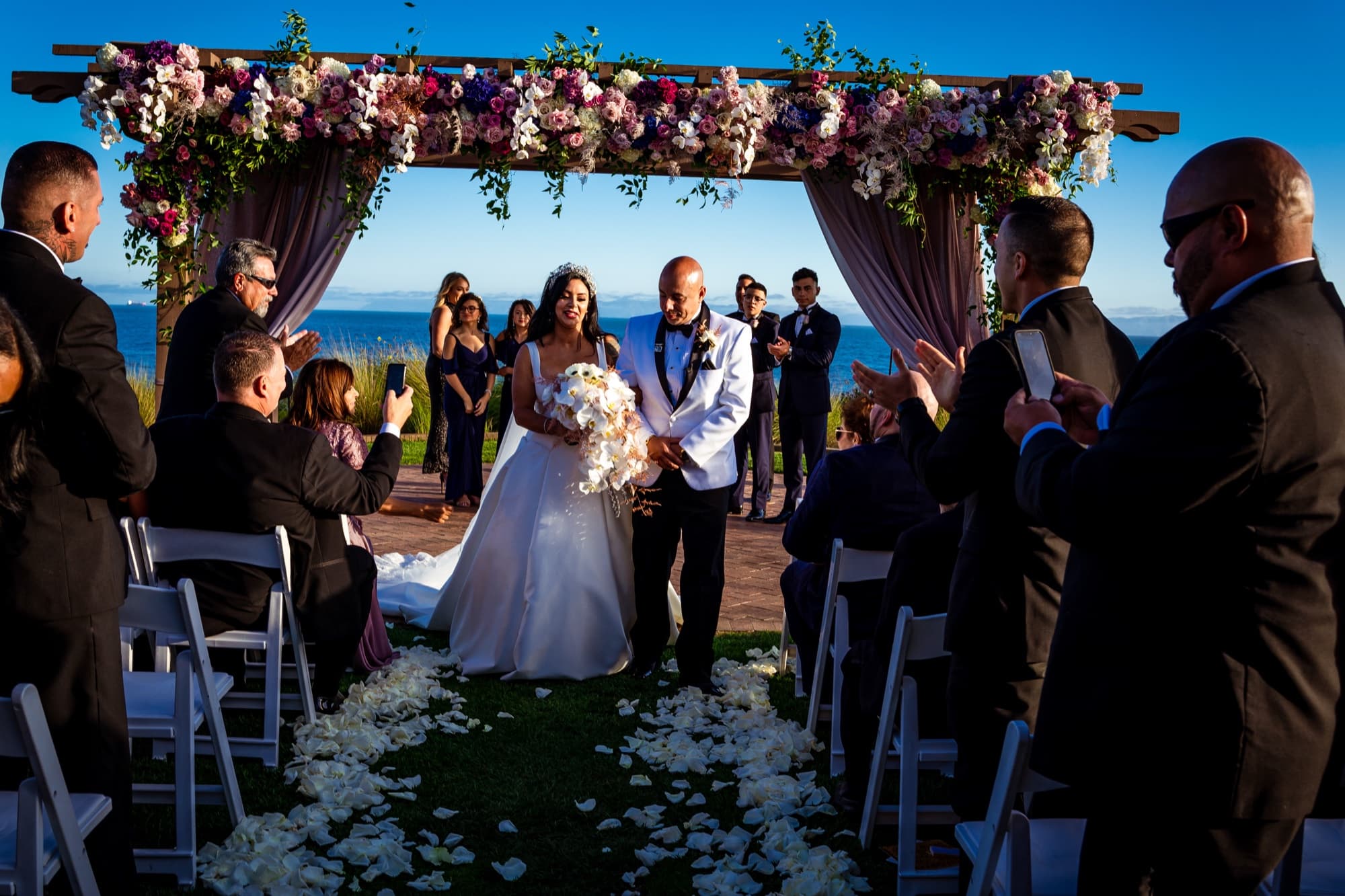 Elise walking the white-petal aisle toward the rose-covered pergola with the Pacific Ocean beyond the bluff at Terranea
