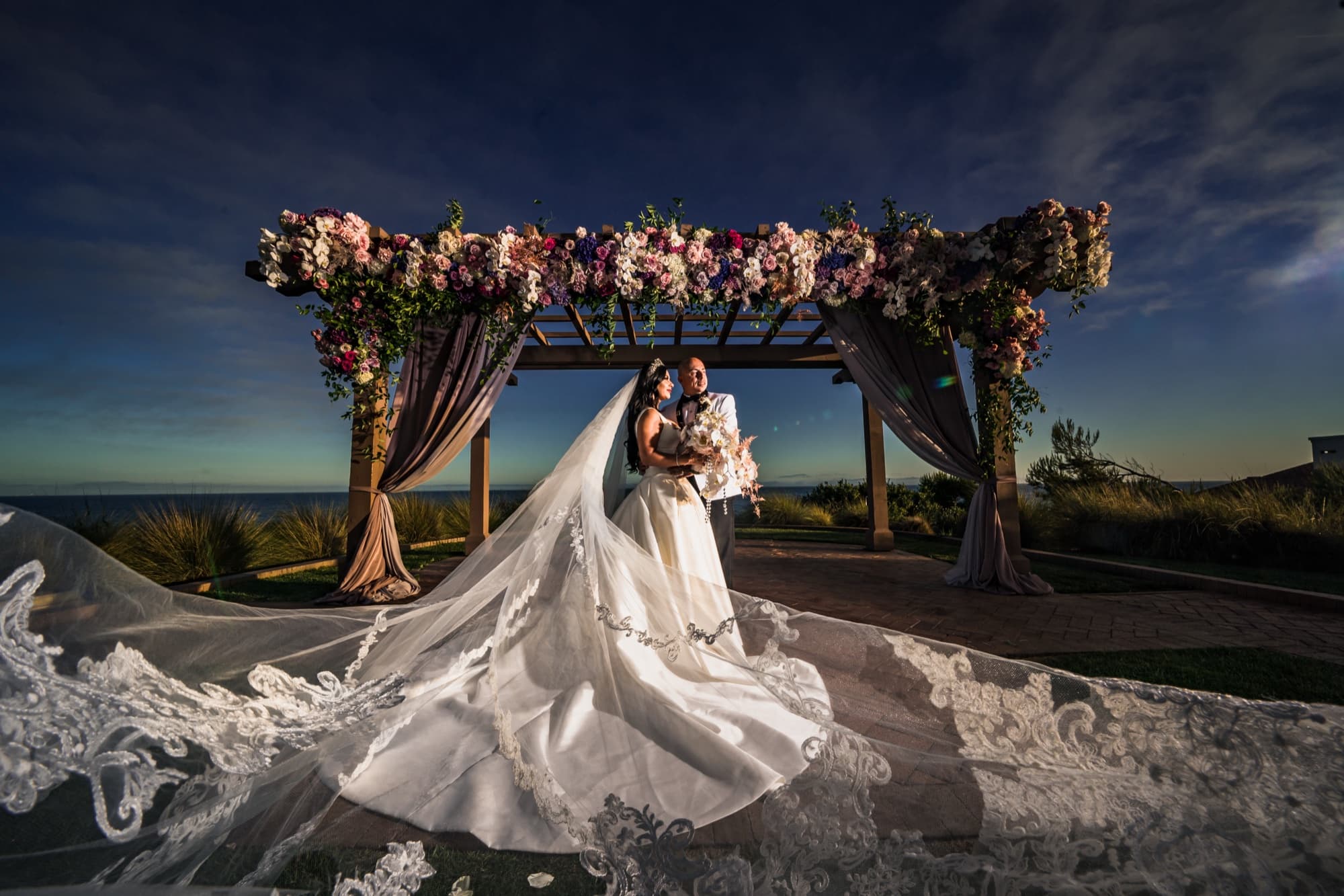 Elise and Luis beneath Terranea's rose-covered ceremony pergola at dusk, her lace cathedral veil spread across the entire foreground, photographed by Michael Anthony Photography