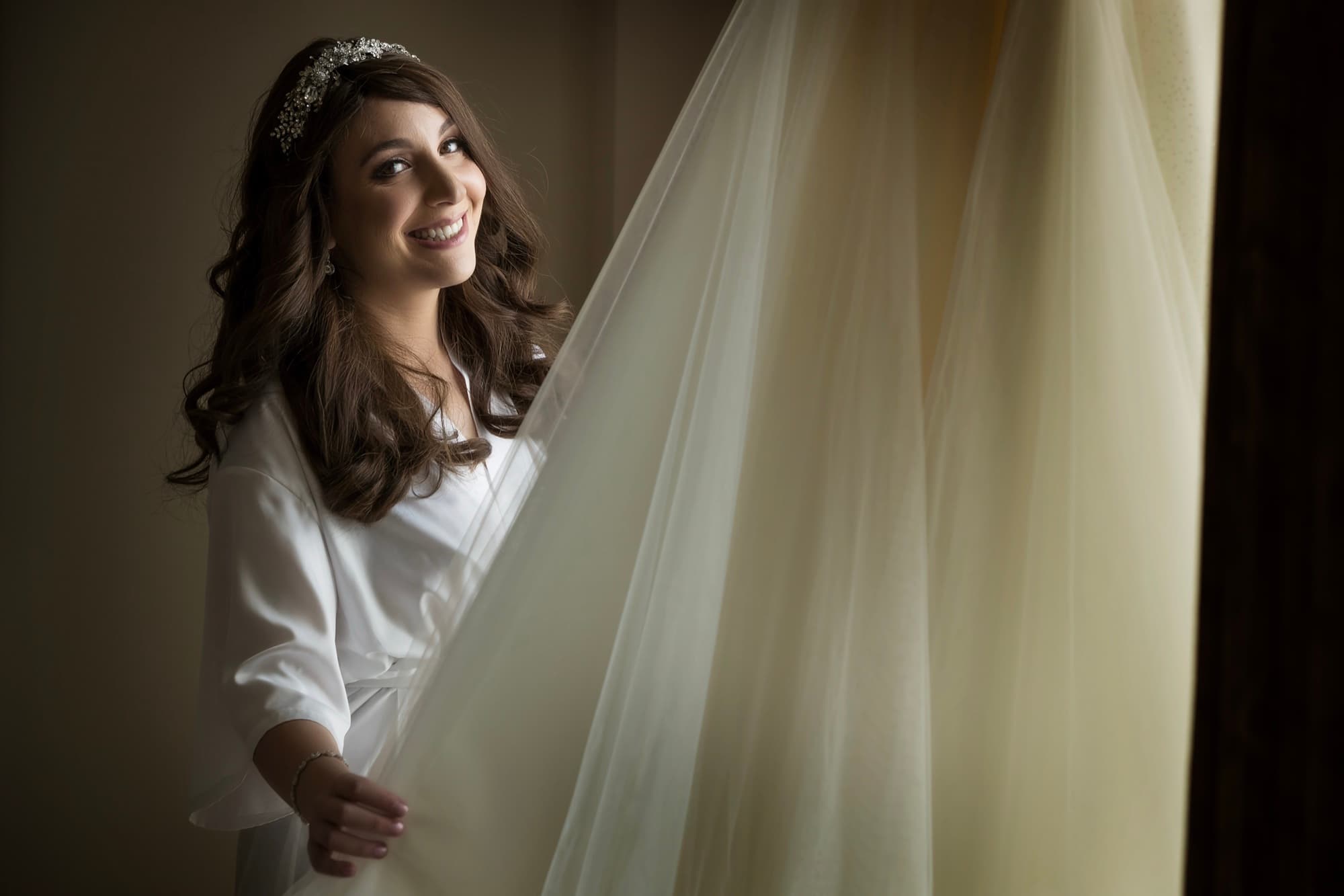 Bride in a white robe smiling beside her hanging veil and gown in the window light of a Terranea Resort suite