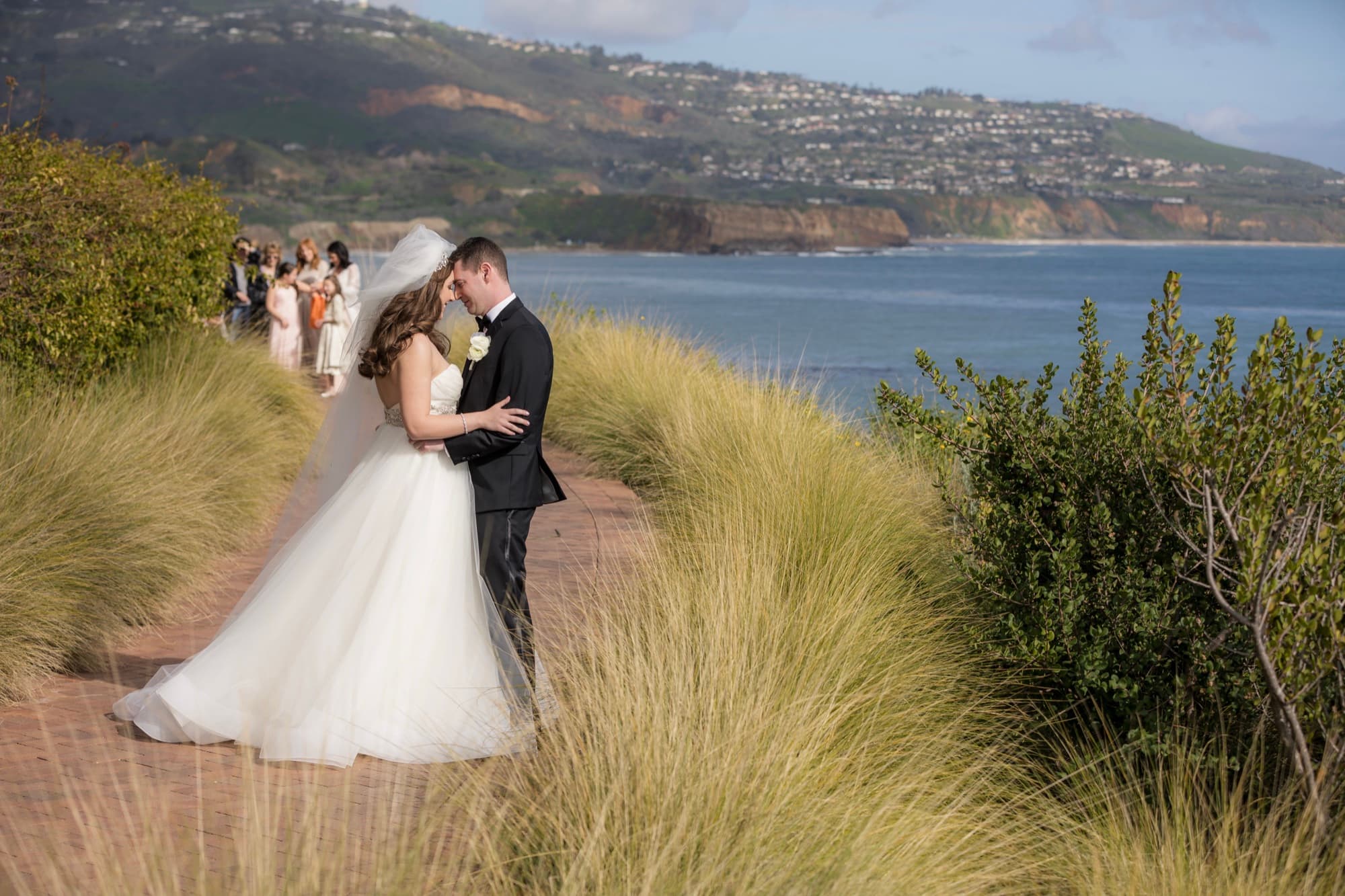 Bride and groom embracing on a bluff-top trail at Terranea Resort with the Palos Verdes coastline stretching behind them