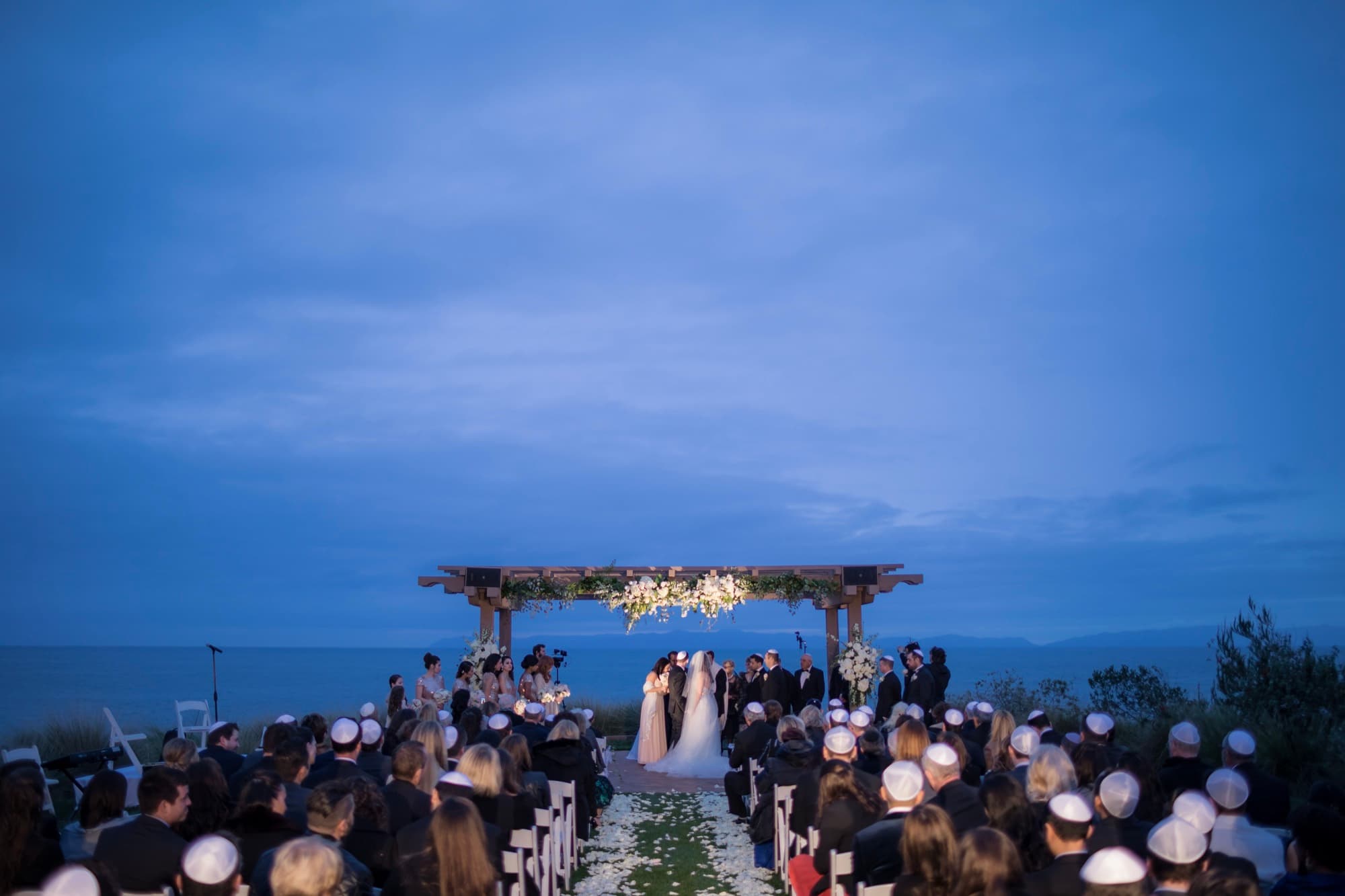 Wide view of a blue-hour chuppah wedding ceremony at Terranea Resort with the Pacific Ocean on the horizon and guests filling the lawn