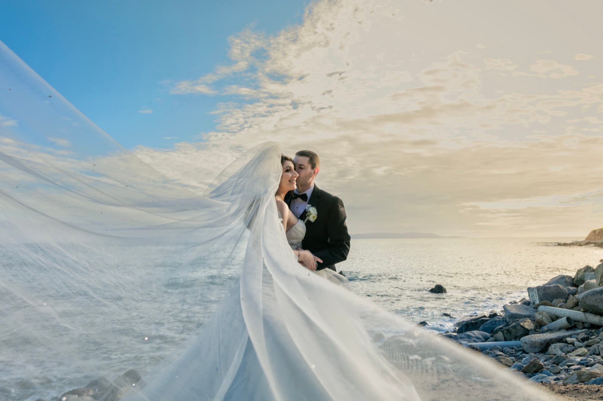 Bride's cathedral veil sweeping across a rocky cove at Terranea Resort at golden hour as the couple embraces by the ocean, photographed by Michael Anthony Photography