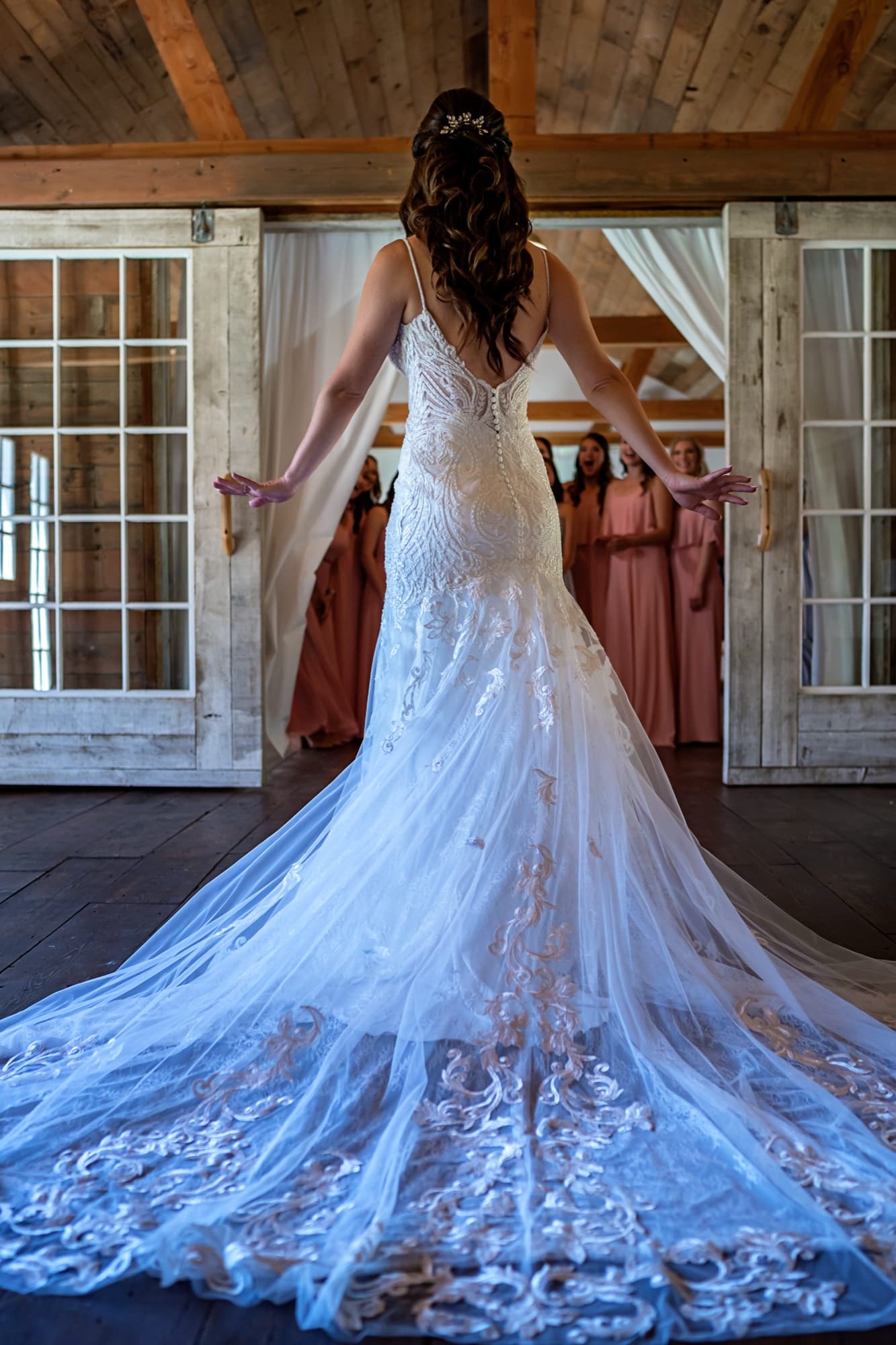 Allison from behind in the barn doorway, her lace train fanned across the floor as her bridesmaids look on in blush and lavender