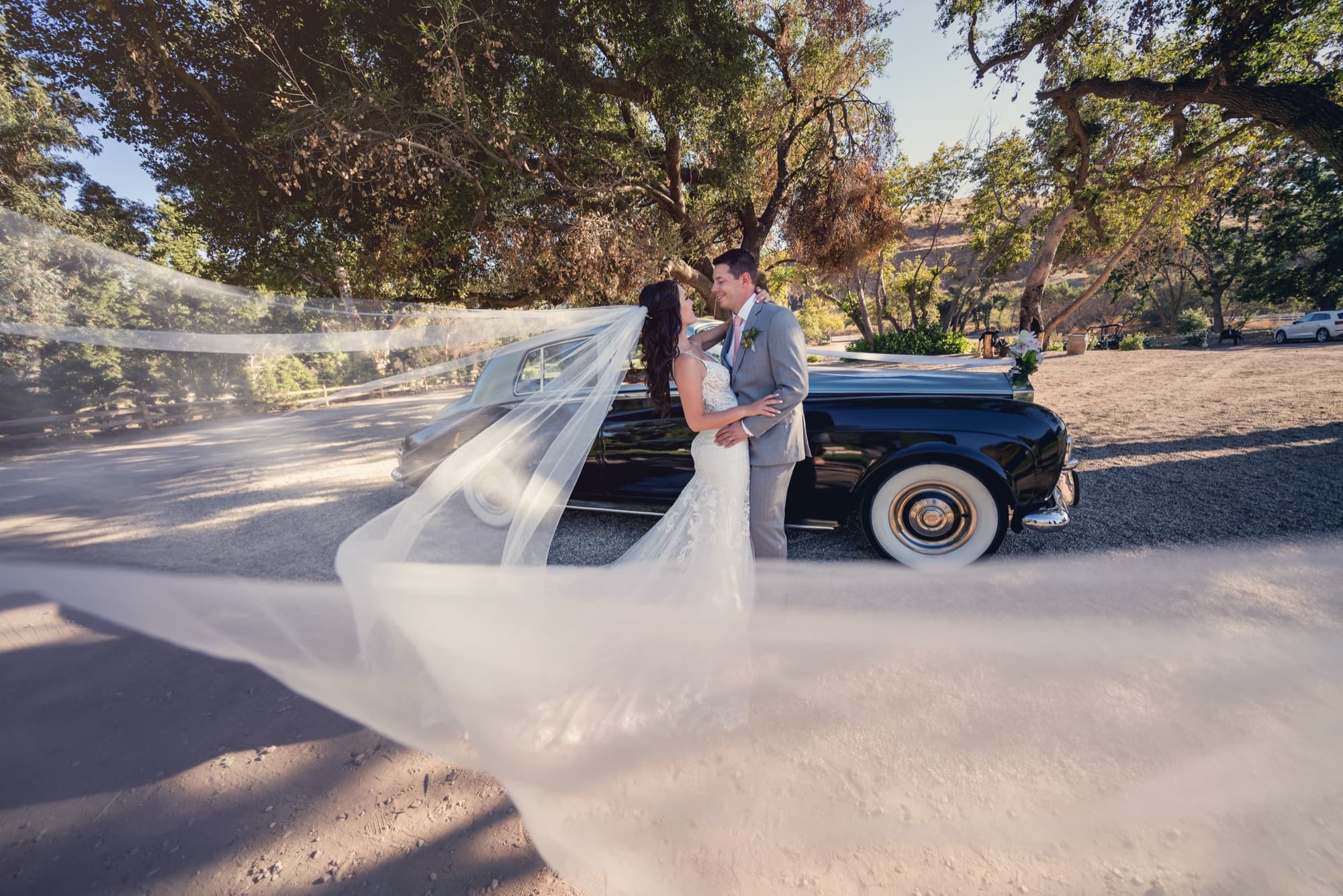 Allison's veil sweeping across the frame past the vintage black car with whitewall tires under the oaks