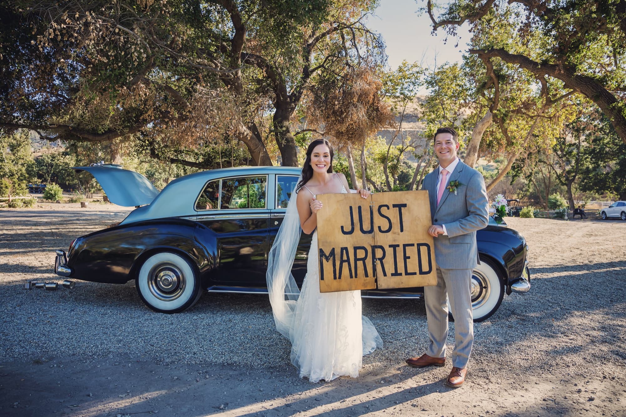 Allison and Joshua holding a hand-lettered Just Married sign beside the vintage car at Triunfo Creek Vineyards