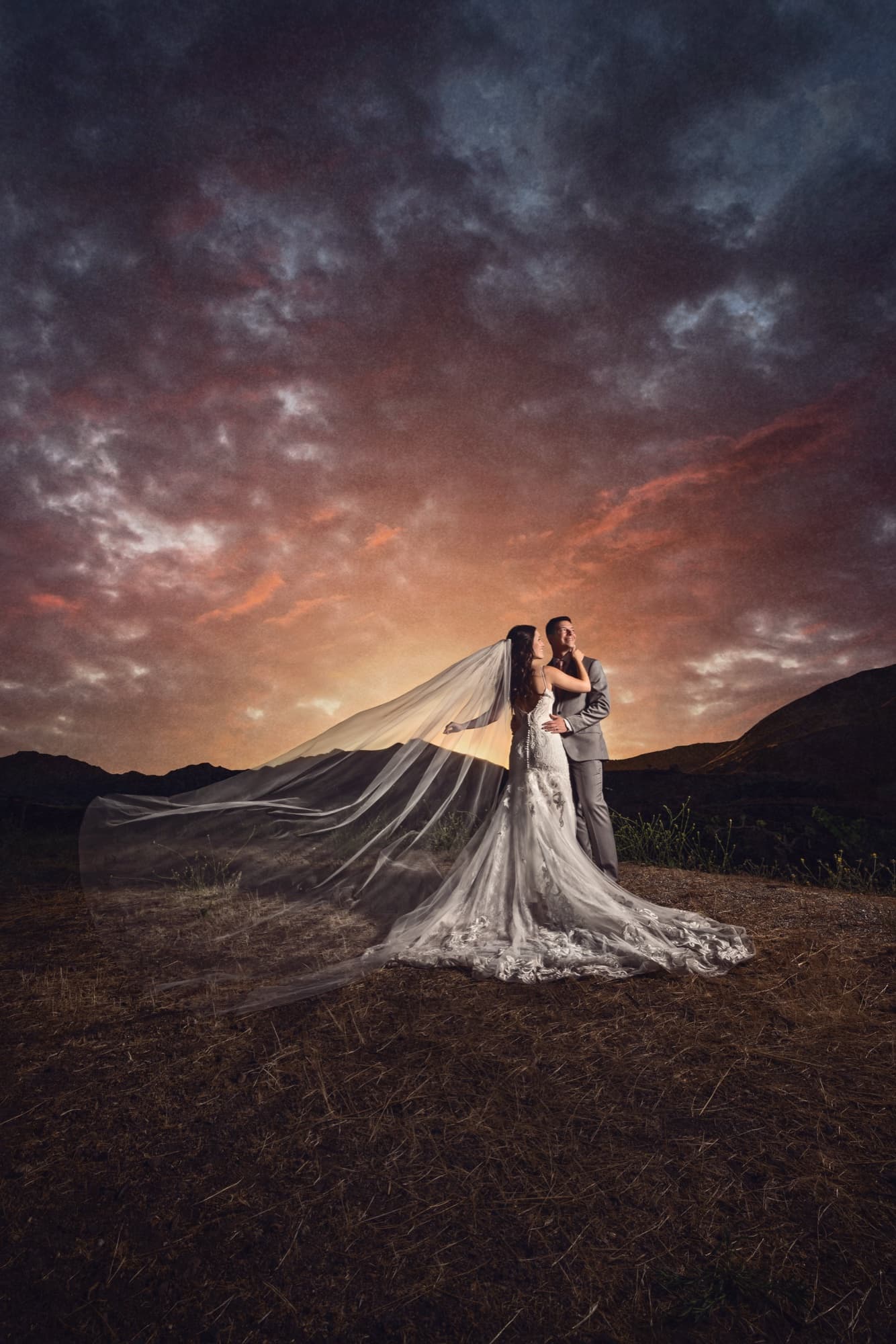 Allison and Joshua embracing on the Triunfo Creek hillside at sunset, her cathedral veil catching the wind beneath a fire-colored sky, photographed by Michael Anthony Photography