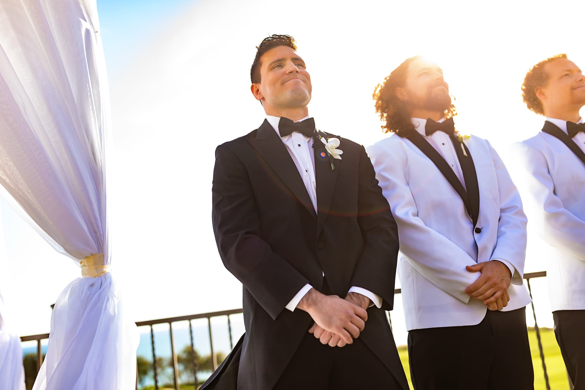 James smiling at the flower-crowned arch with sun flare behind him as he watches Jamie walk down the aisle