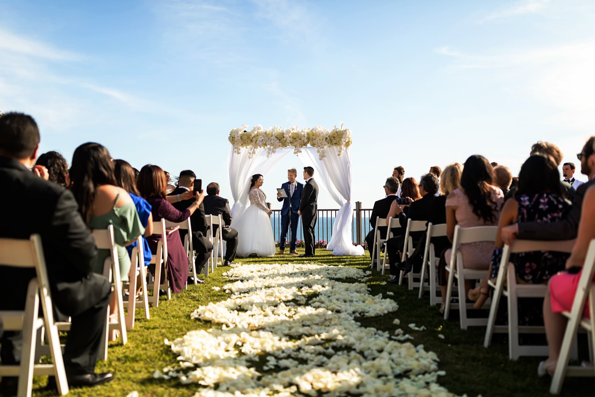 Wide view of the clifftop ceremony at Trump National Golf Club, a white petal-lined aisle leading to the orchid arch above the Pacific