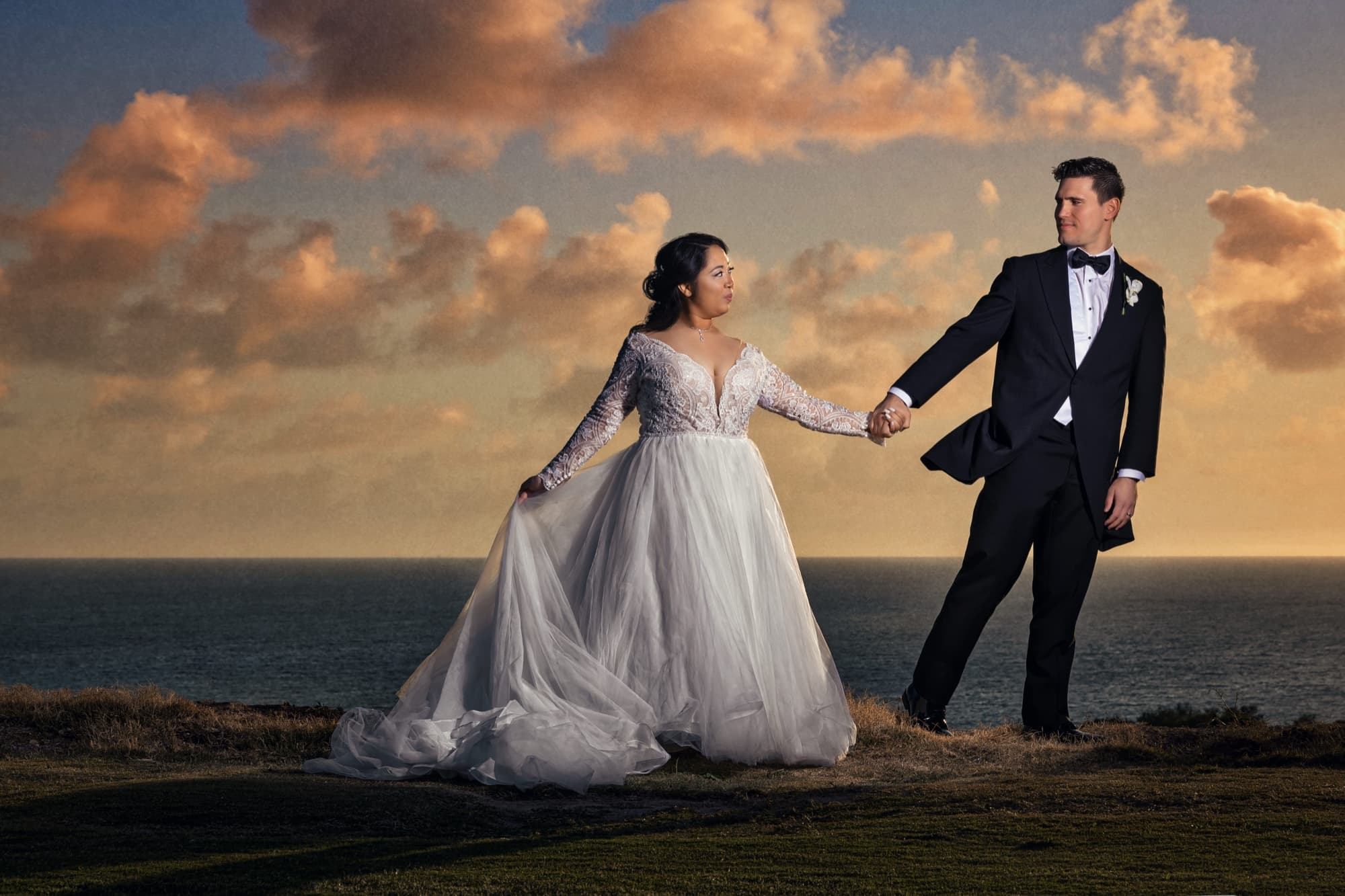 Jamie and James walking hand in hand along the clifftop at Trump National Golf Club at sunset, her tulle gown catching the wind beneath pink storm clouds over the Pacific, photographed by Michael Anthony Photography
