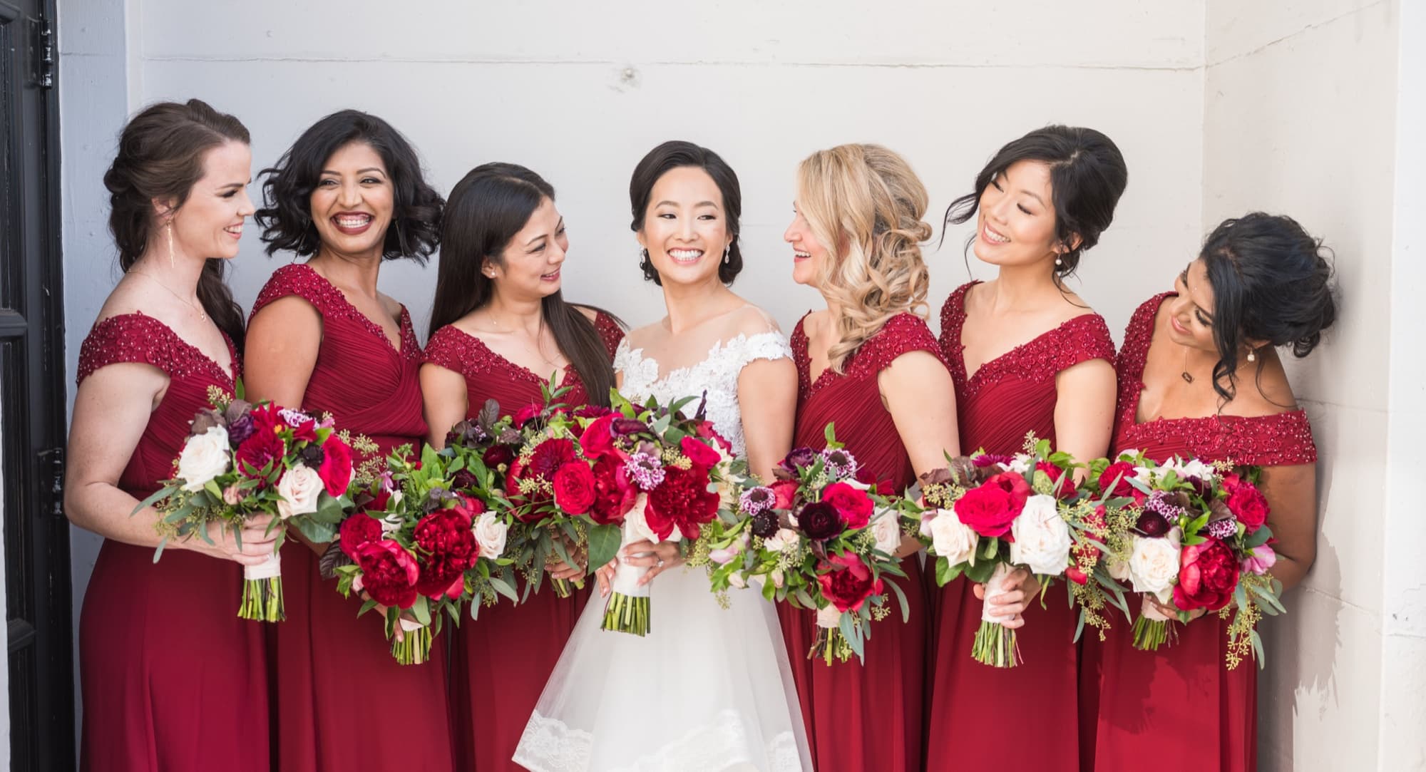 Bride laughing with six bridesmaids in deep red gowns holding burgundy and ivory bouquets at Vibiana