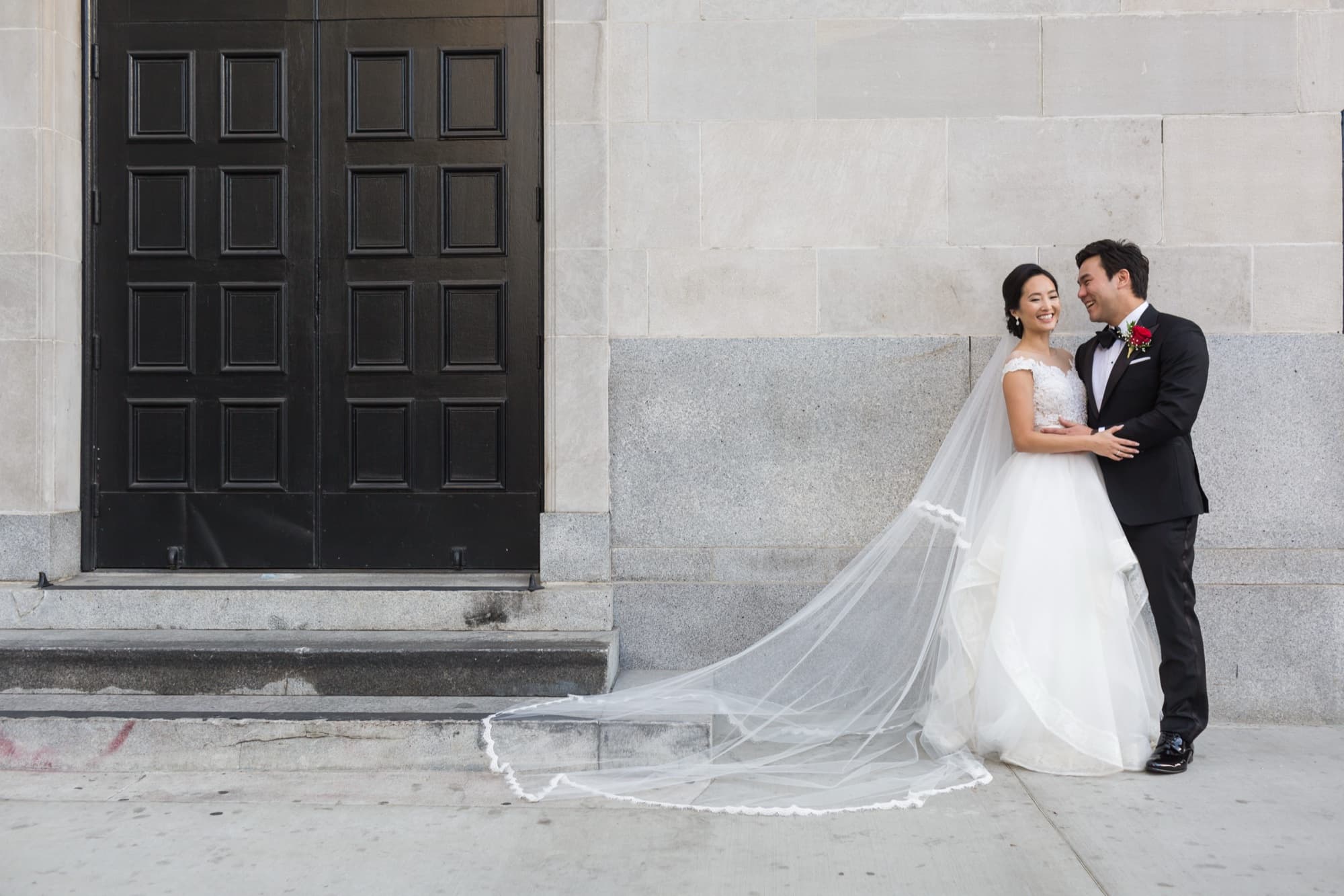 Bride and groom embracing beneath Vibiana's monumental stone facade at dusk with the cathedral veil sweeping across the steps
