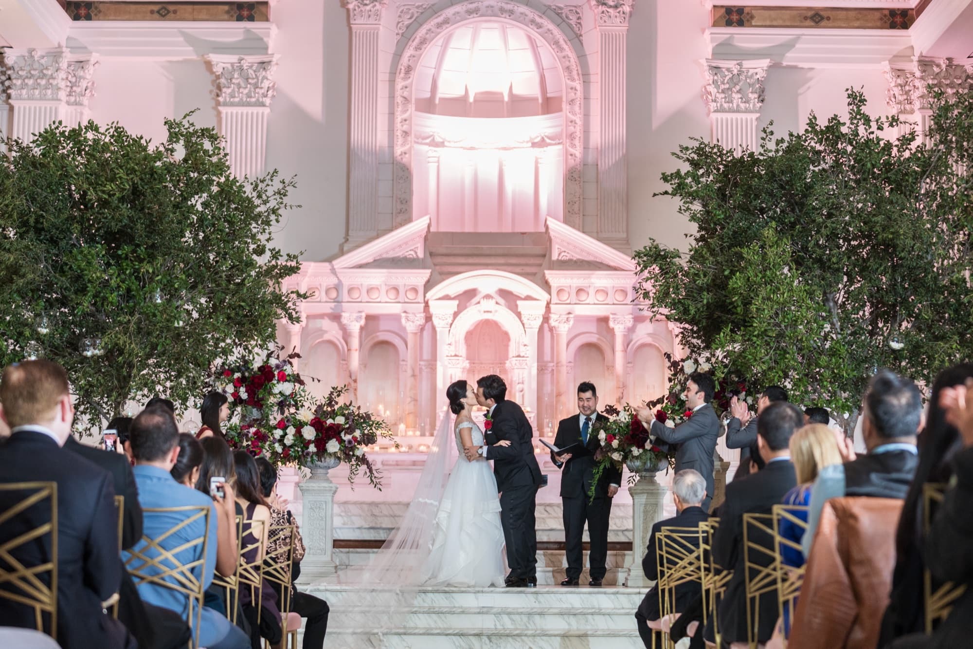 Wedding ceremony at the pink-lit marble altar of Vibiana's main hall with olive trees and red rose arrangements flanking the couple