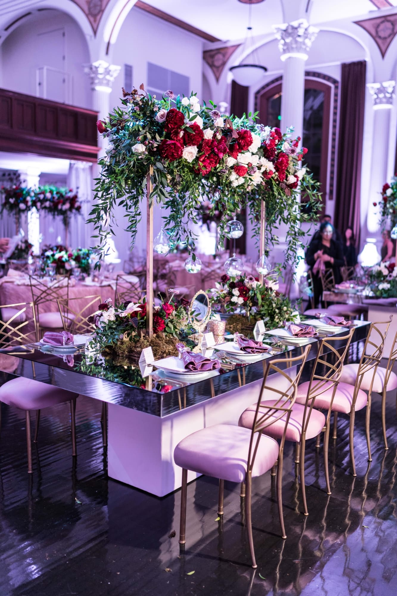 Mirrored reception table at Vibiana with gold chairs, blush linens, and a tall centerpiece of burgundy and ivory flowers under violet uplighting
