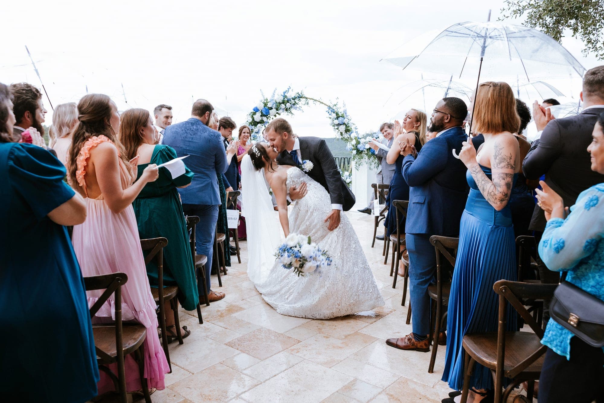 A dip kiss in the aisle among cheering guests holding clear umbrellas