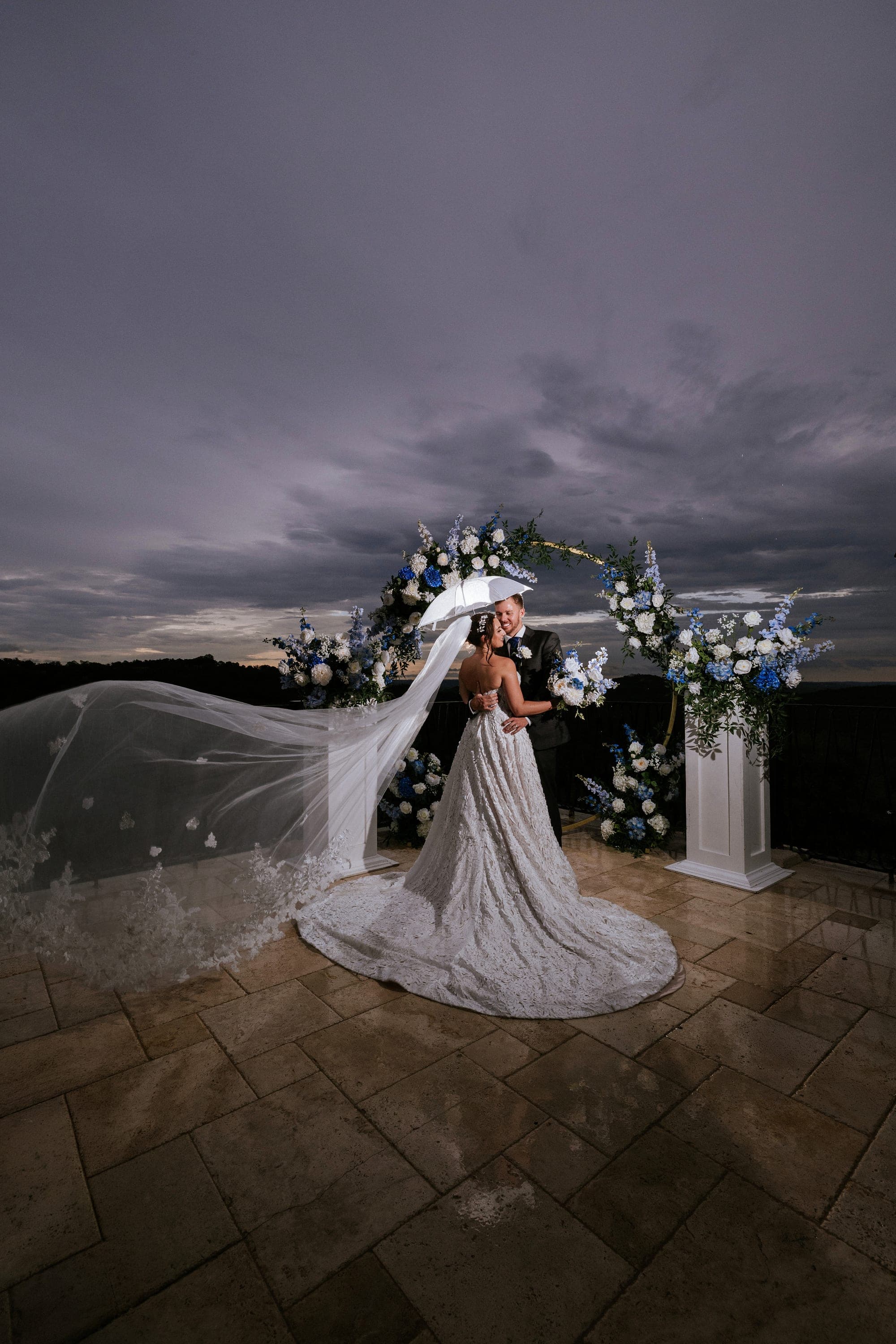 Kelsey and Eric beneath a white umbrella at the arch under a slate storm sky, her veil spread across the wet stone