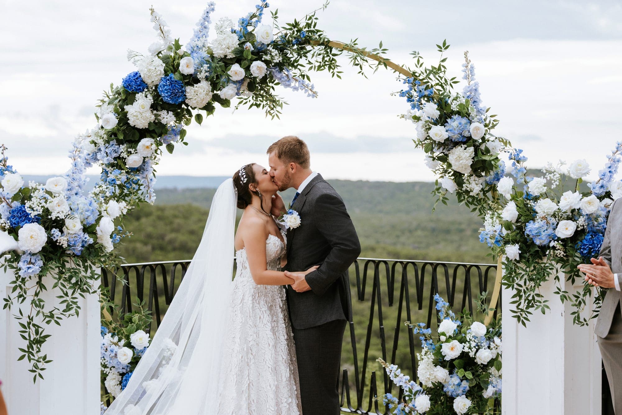 The first kiss beneath the gold hoop arch of blue and white florals, the Hill Country behind