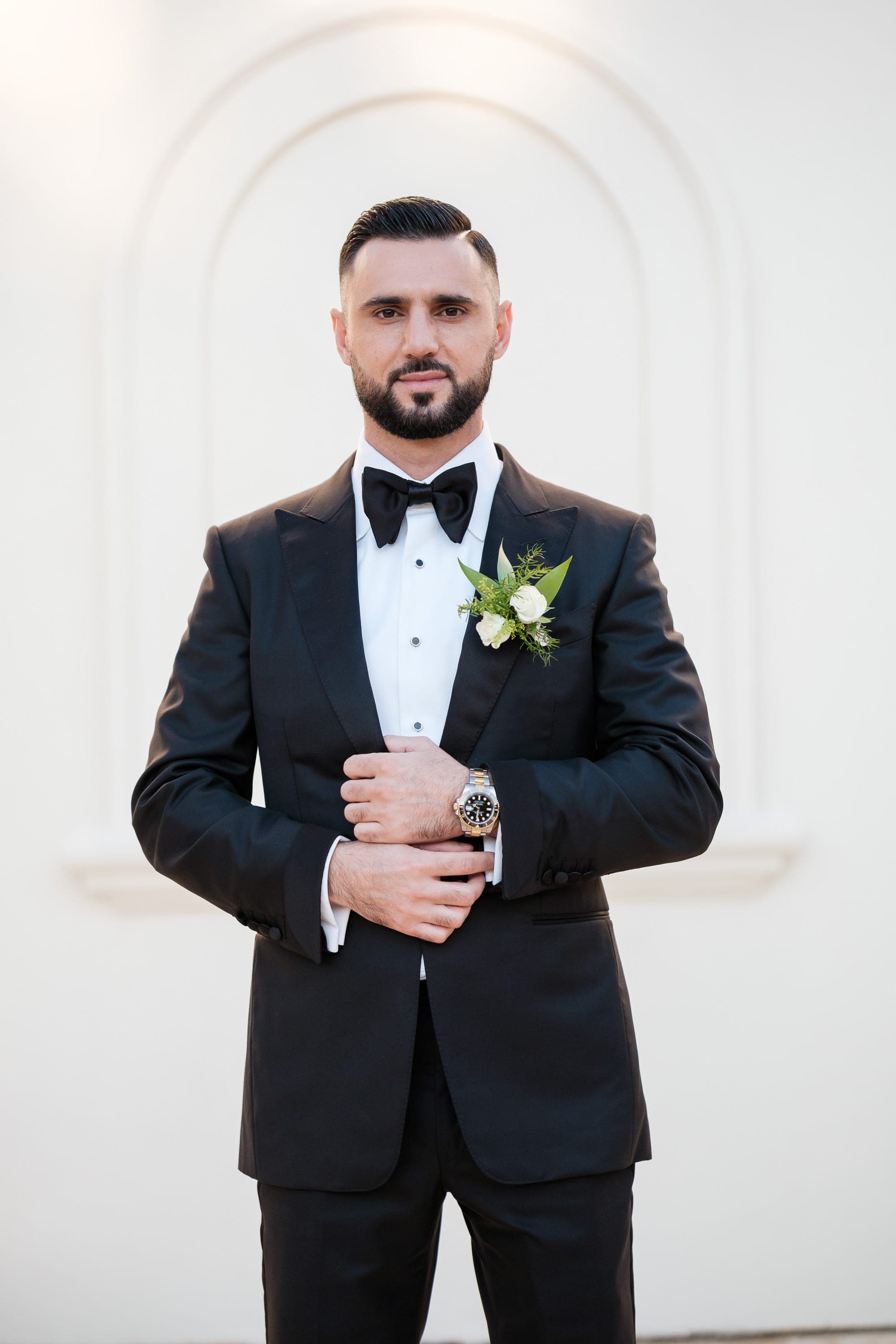 A groom in a black tuxedo and bow tie adjusts his cufflink before a white arched wall.