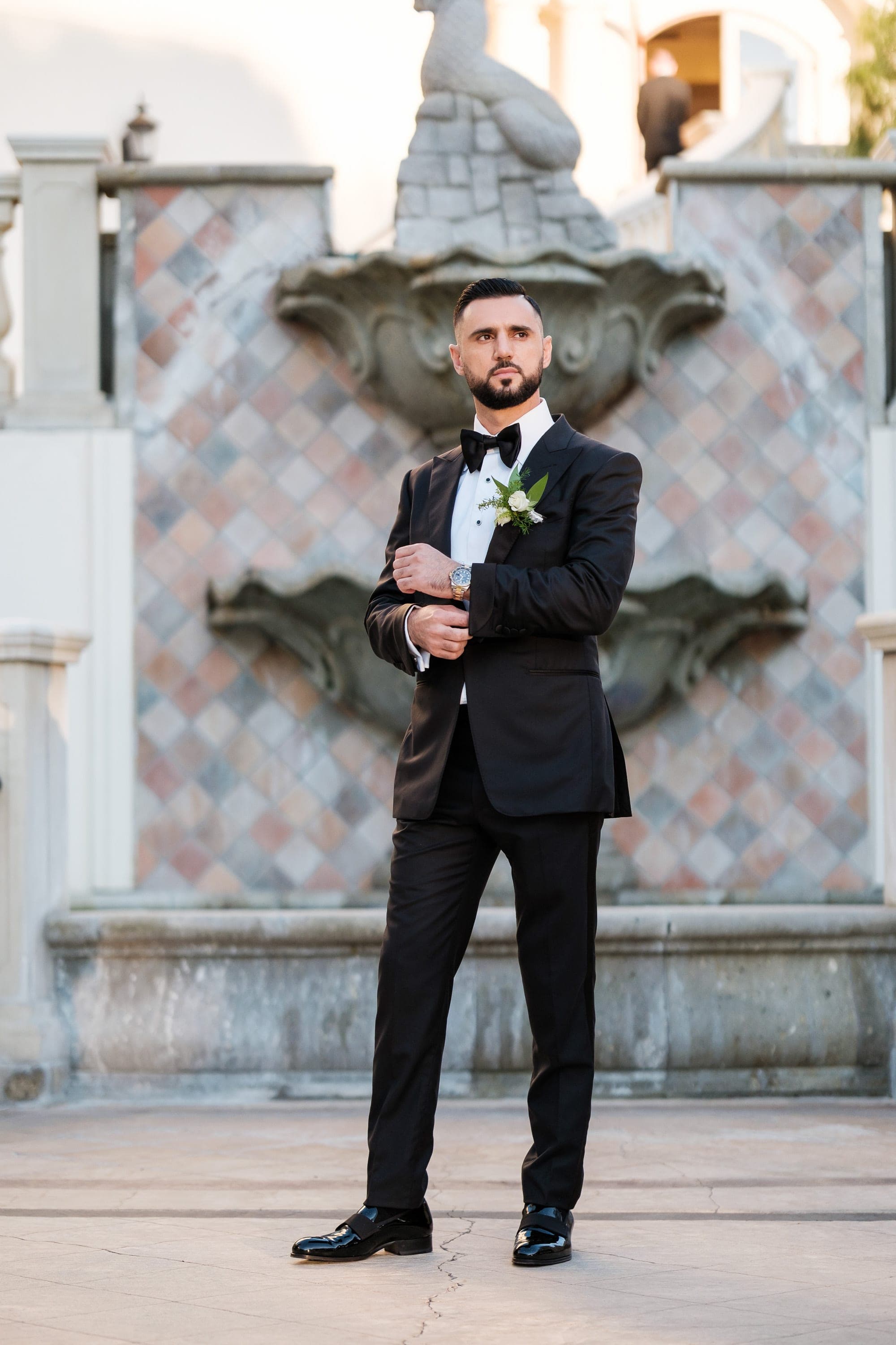 Full-length shot of a groom in a black tuxedo standing before a tiled fountain at golden hour.