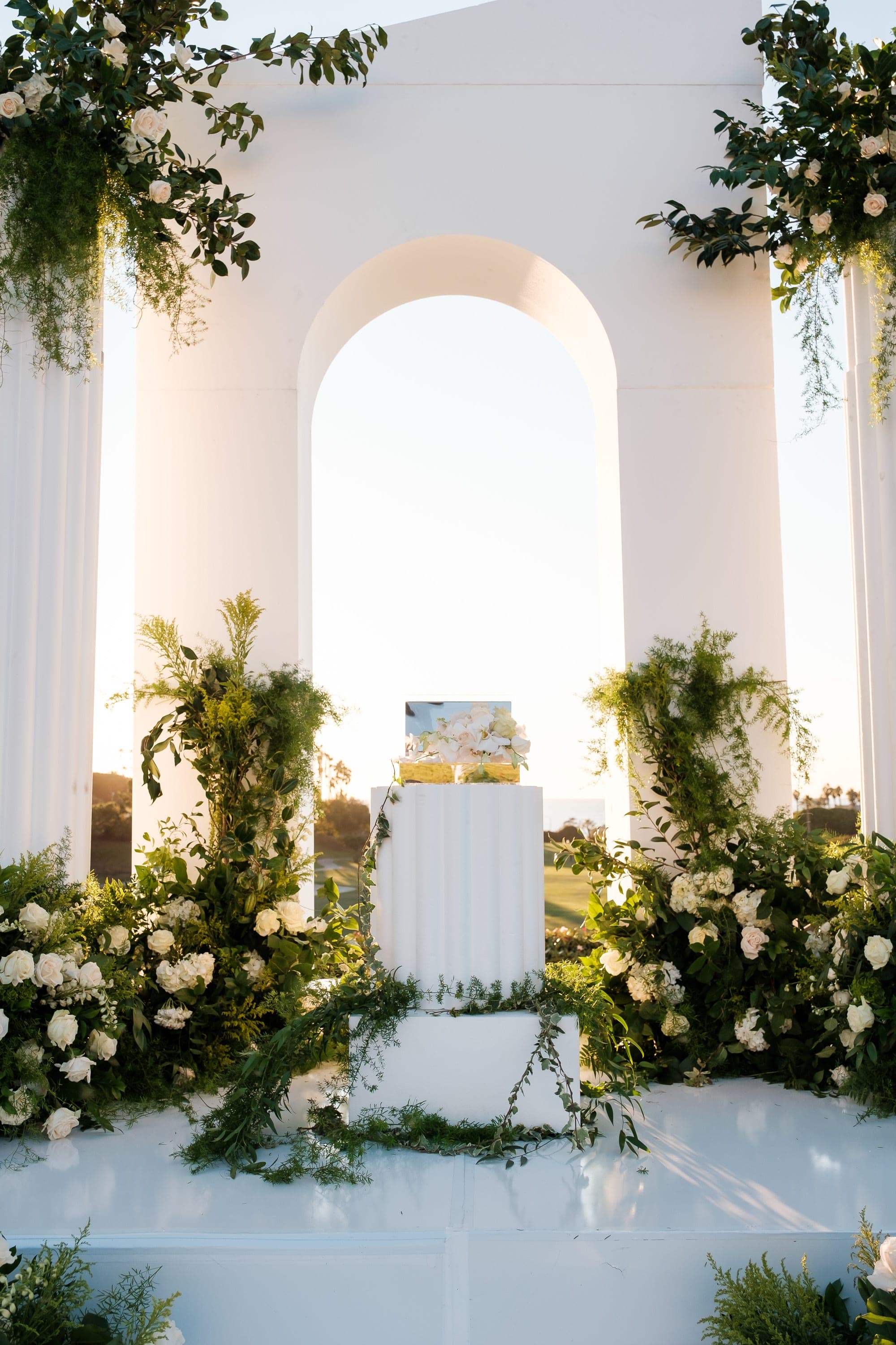 A white floral and greenery arch frames a pedestal holding a small cake at sunset.