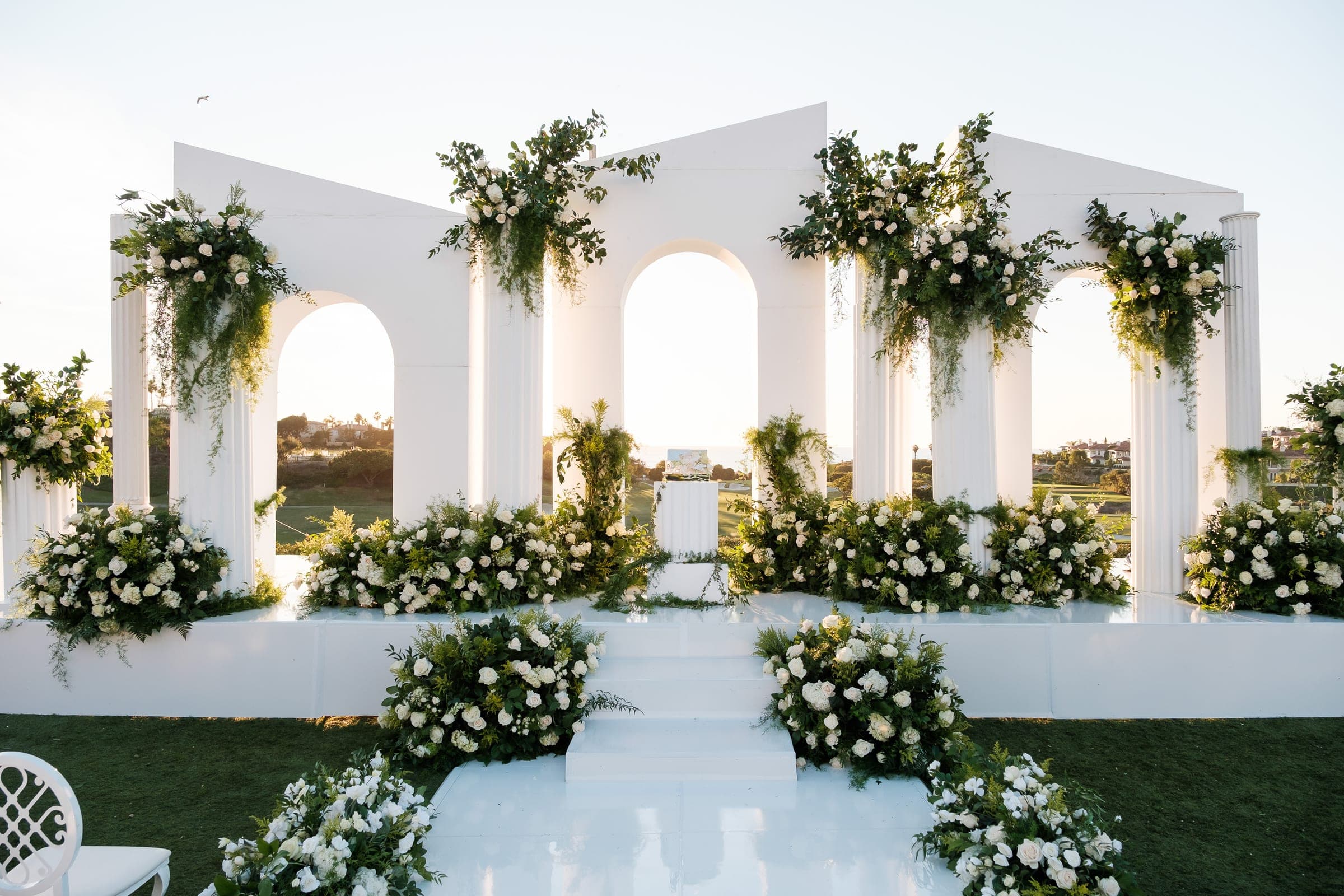 An elaborate white ceremony stage with columns, arches, and abundant white roses and greenery.
