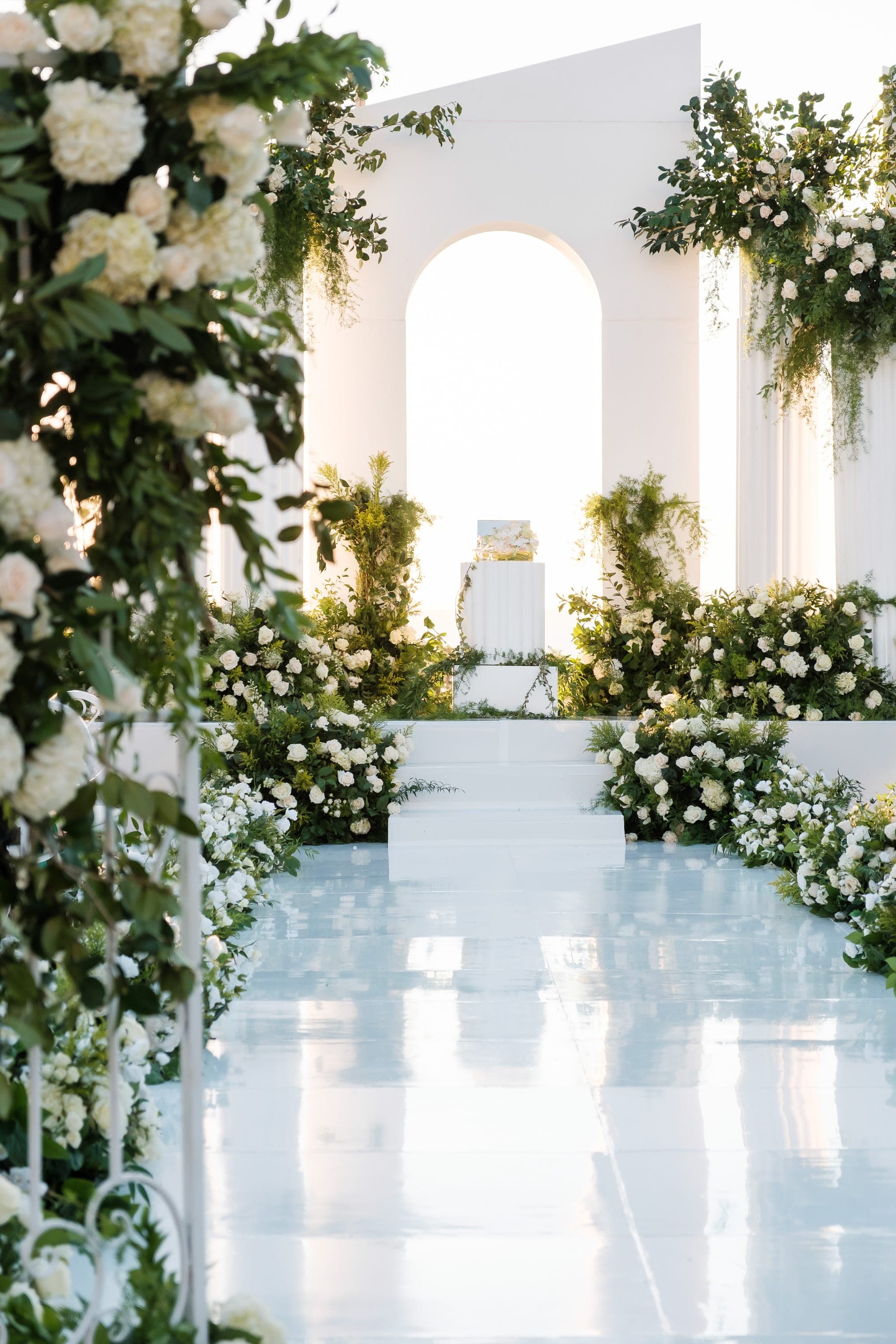 White roses and greenery line a glossy aisle leading toward the arched ceremony backdrop.