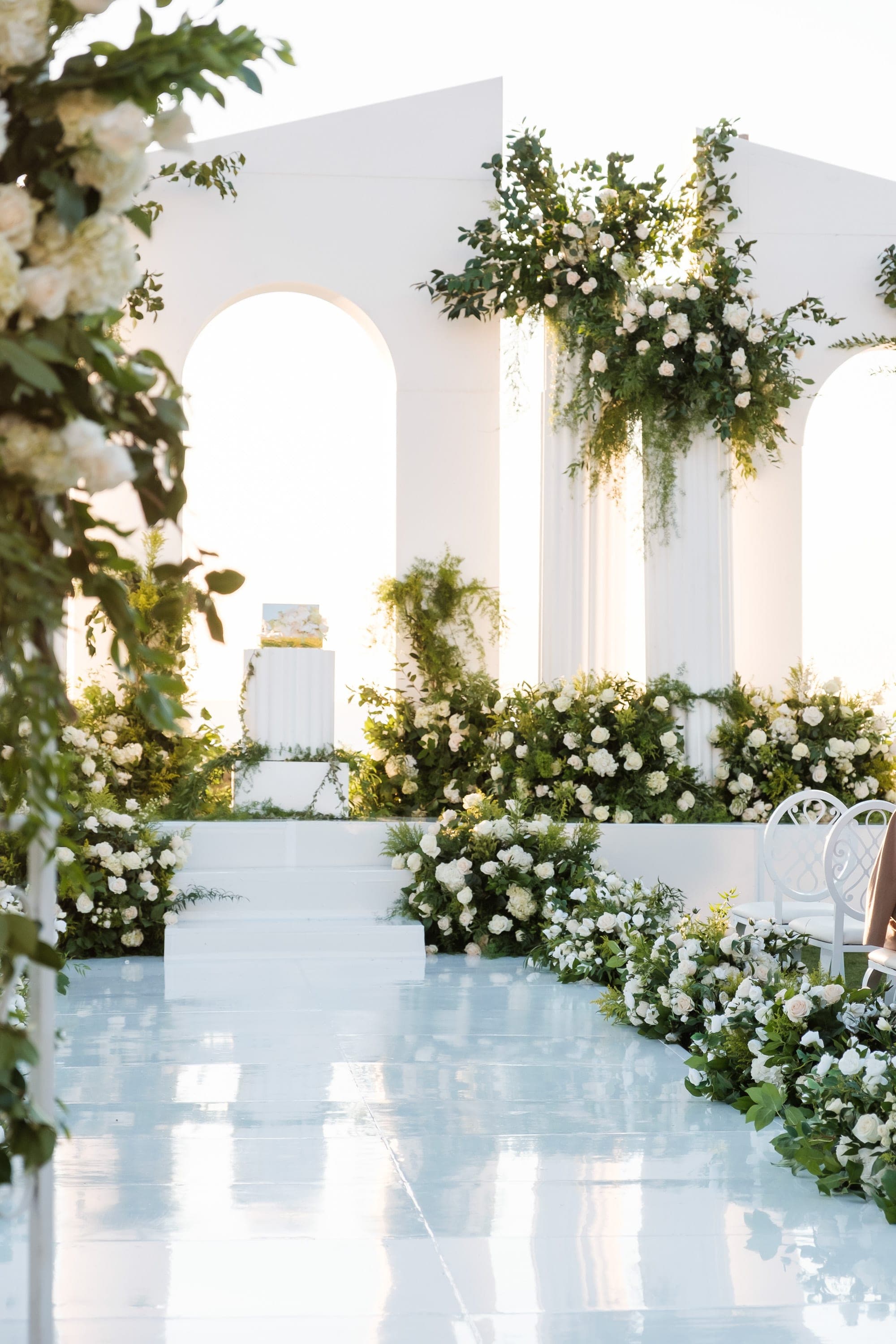Lush white roses and greenery decorate the ceremony steps beside an elegant white chair in warm light.