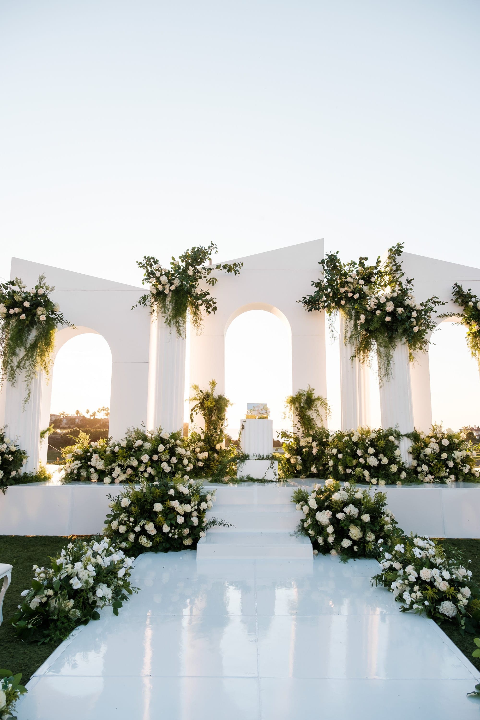 A white aisle leads to arched columns draped in greenery and abundant white floral arrangements at dusk.