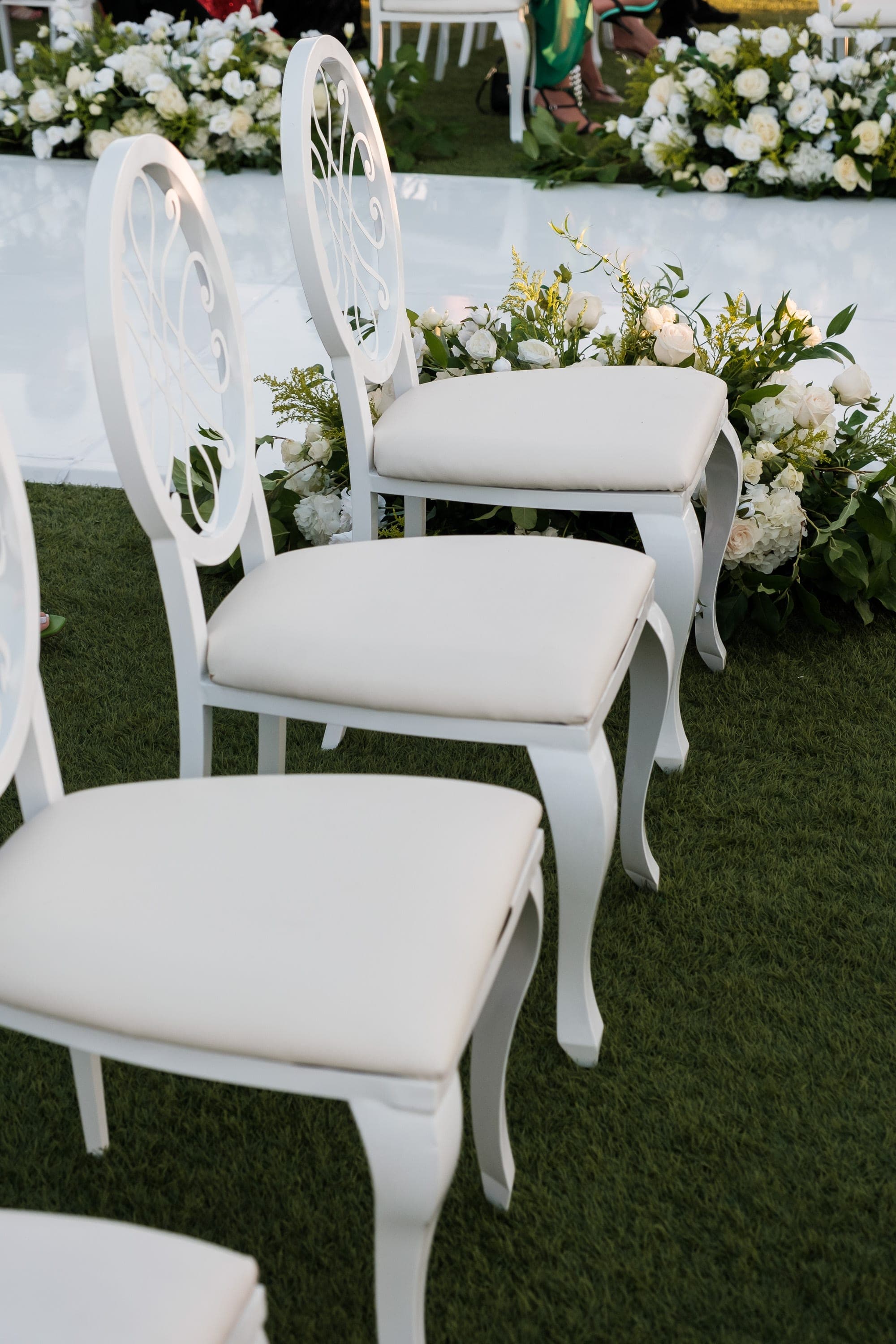 Elegant white chairs with ornate scrolled backs sit on grass beside white roses and greenery.