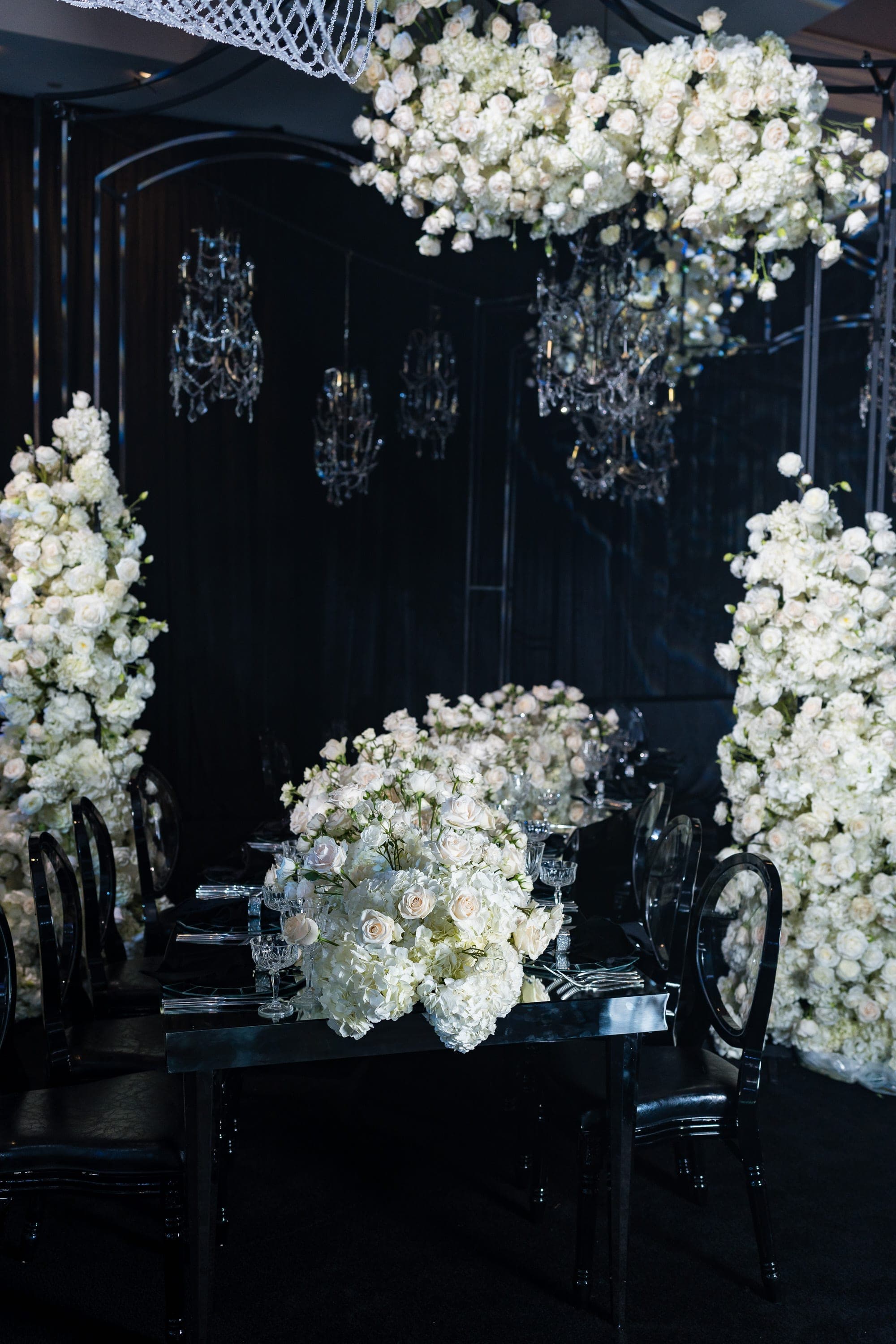 White roses and hydrangeas surround a black mirrored dining table lit by crystal chandeliers.