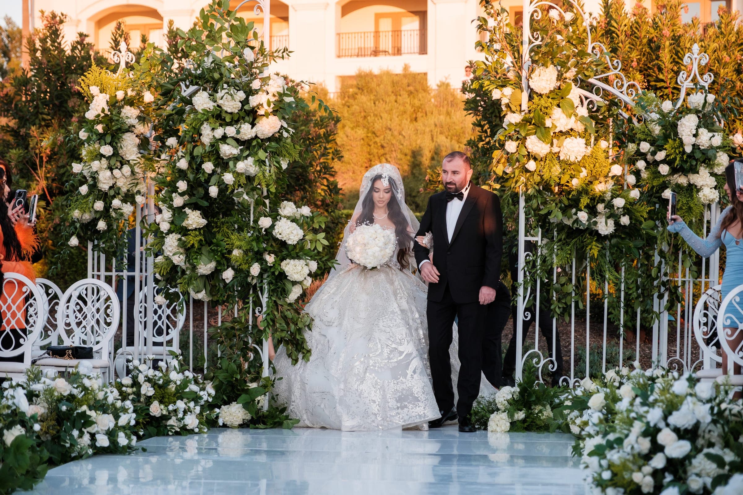 A bride and her father walk the aisle between towering white floral gates in warm light.