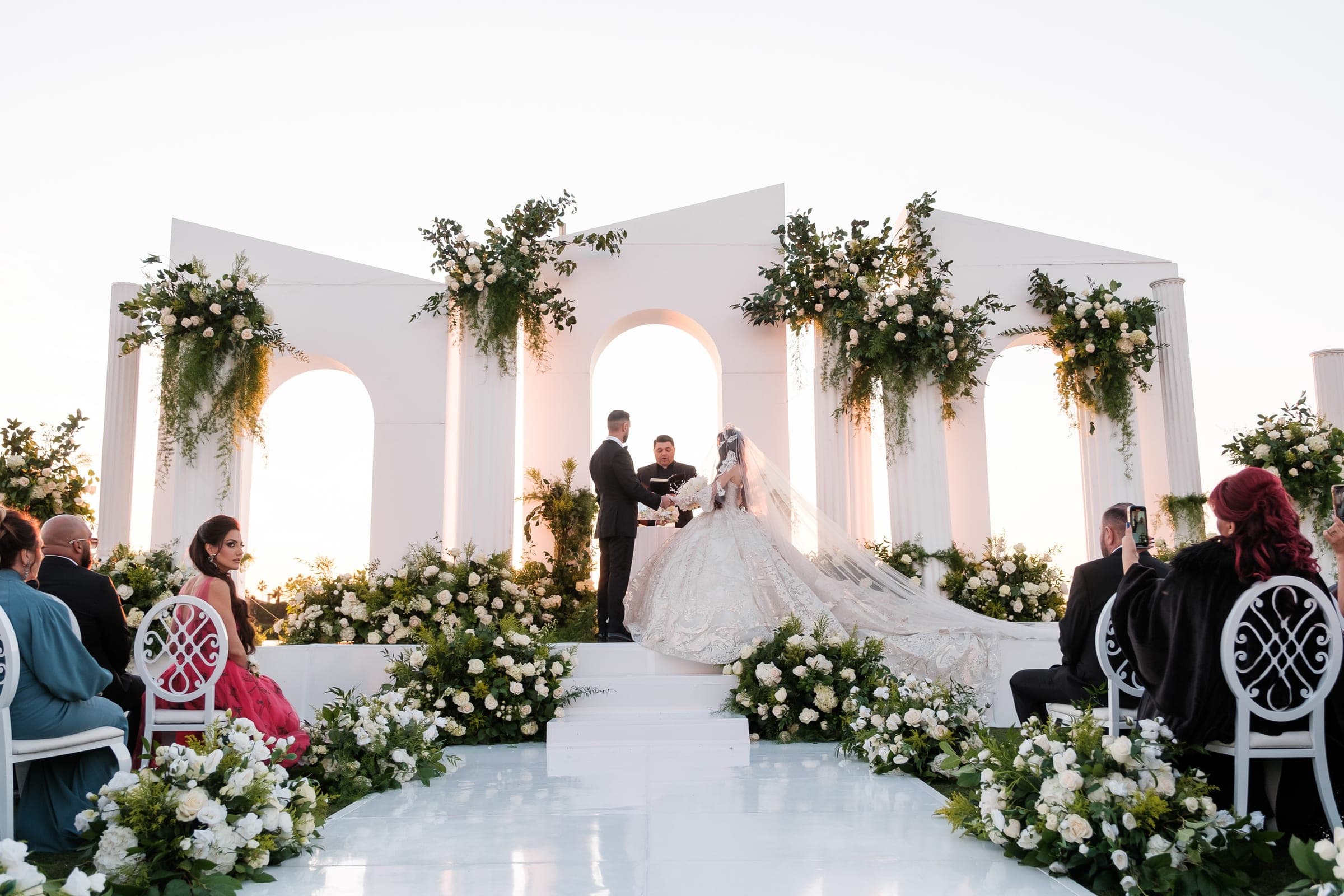 Outdoor sunset ceremony with bride and groom before white arches, columns, and lush greenery amid seated guests