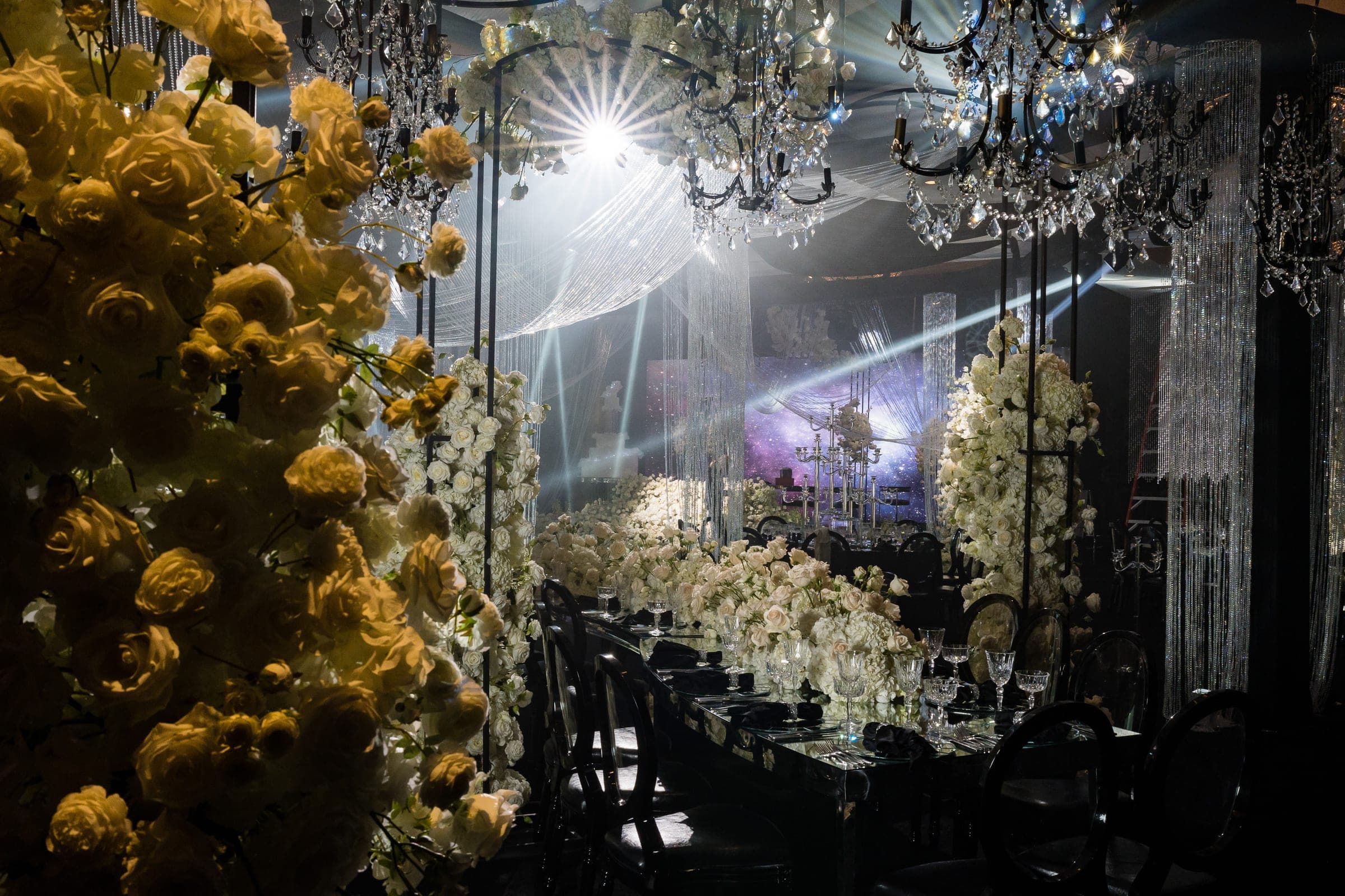 Dim reception hall with cascading yellow and white roses, crystal chandeliers, and bright stage lights beyond