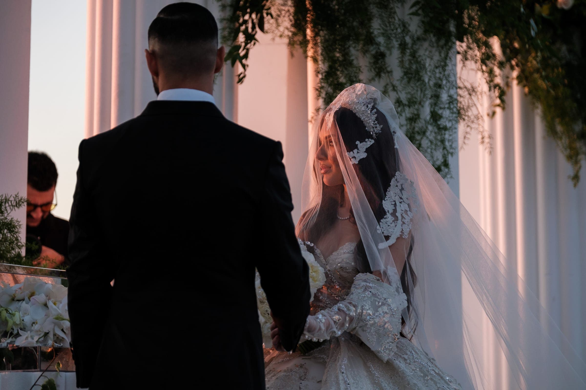 Bride in an ornate veil faces her suited groom near glowing white columns at dusk