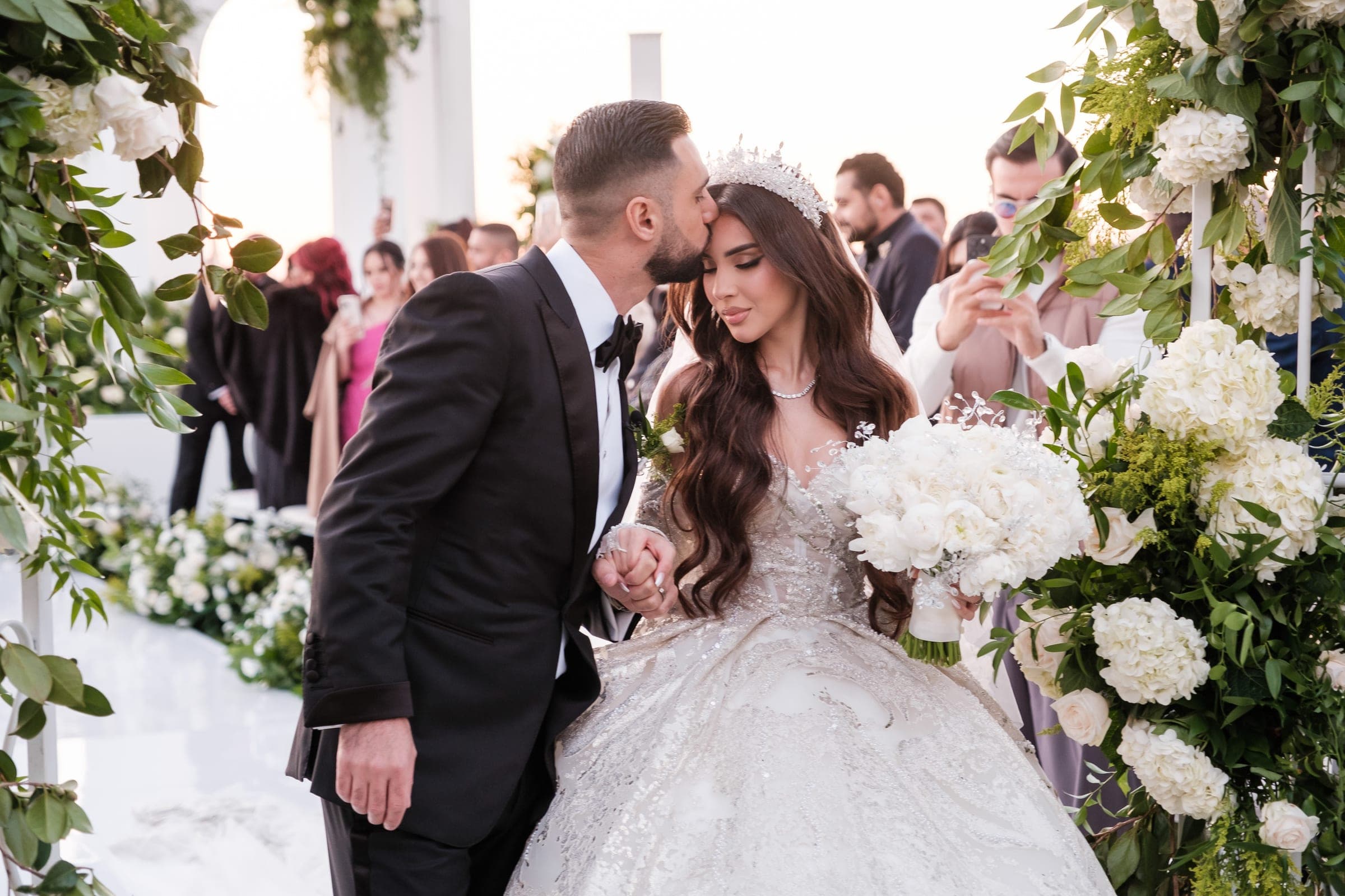 Groom kisses his crowned bride on the cheek among white florals as guests watch and film