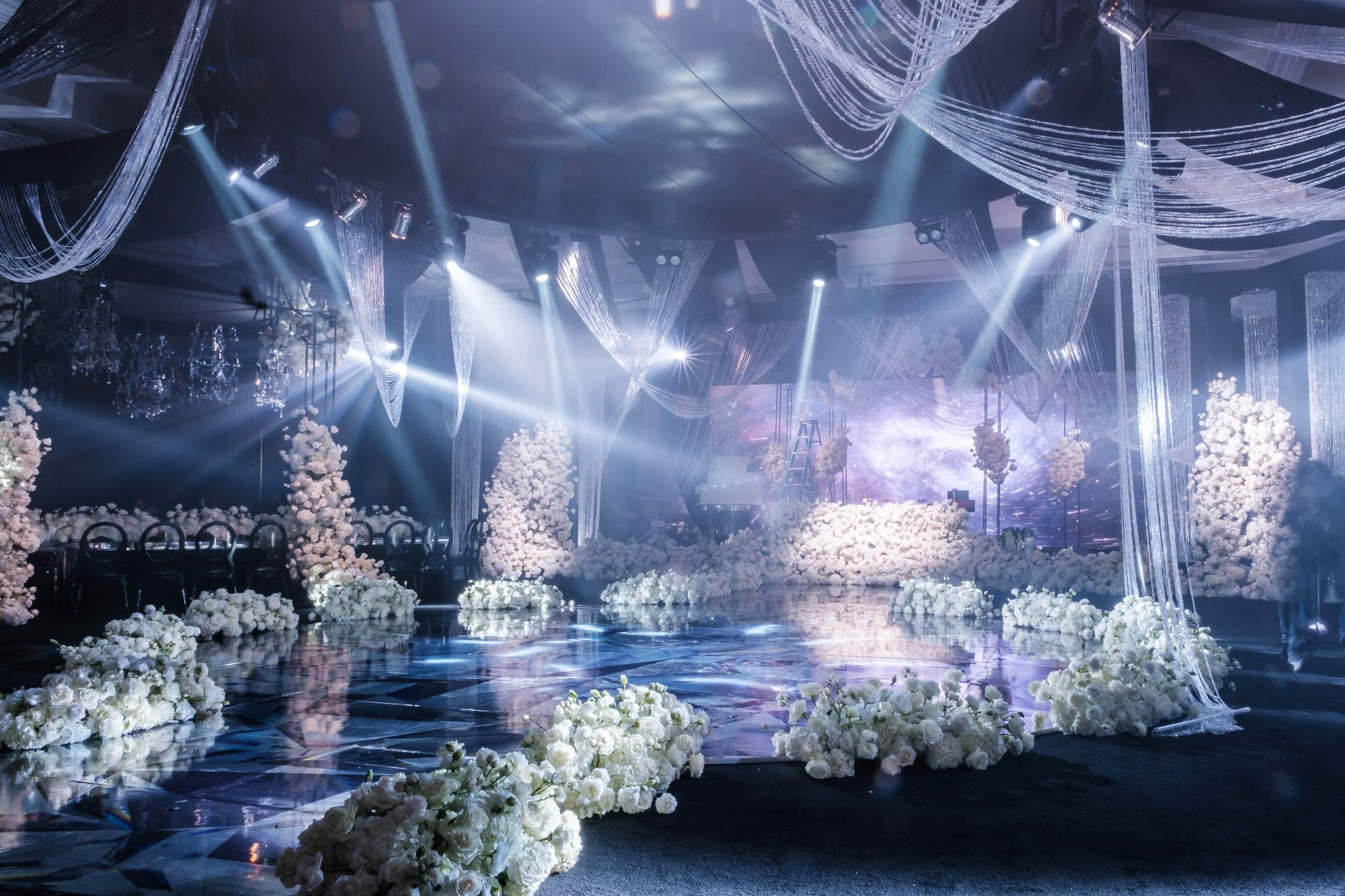 Dramatic ballroom with white rose arrangements, hanging beaded strands, and dance floor washed in blue spotlights