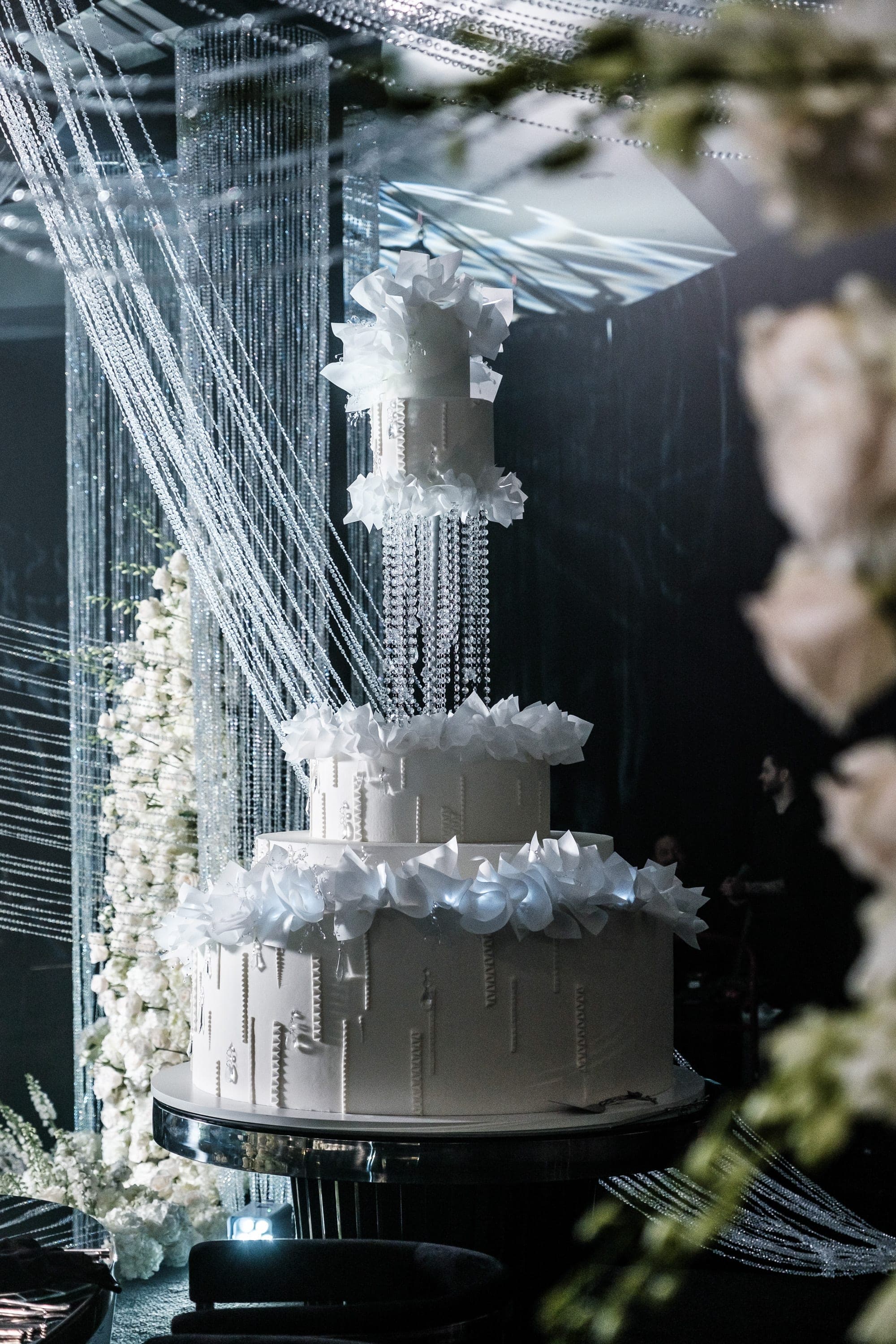 Towering white wedding cake adorned with sugar flowers and crystal strands beside cascading beaded curtains and roses.