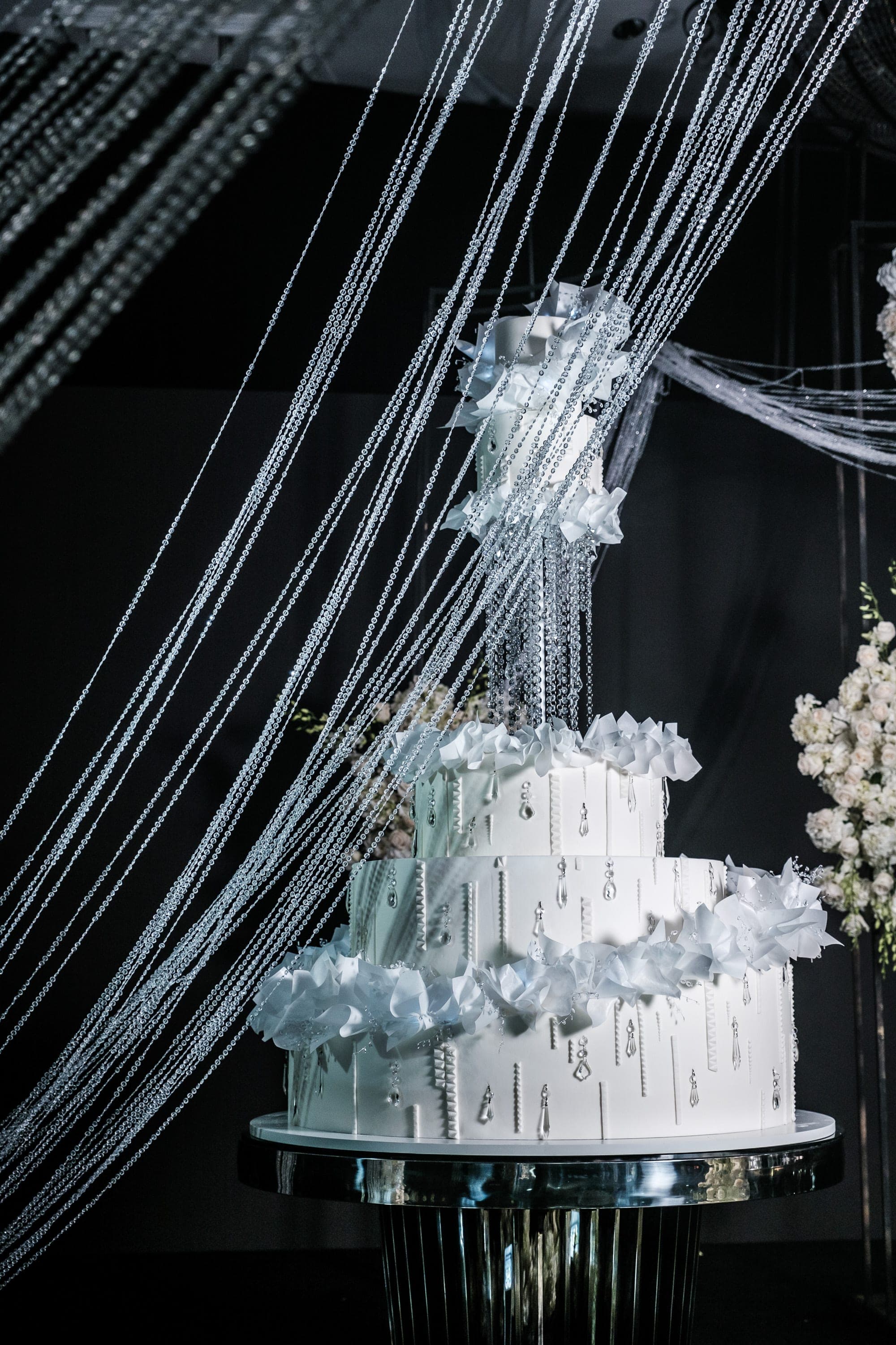 Multi-tier white cake with ruffled sugar flowers and hanging crystals against dark draped beaded backdrop.