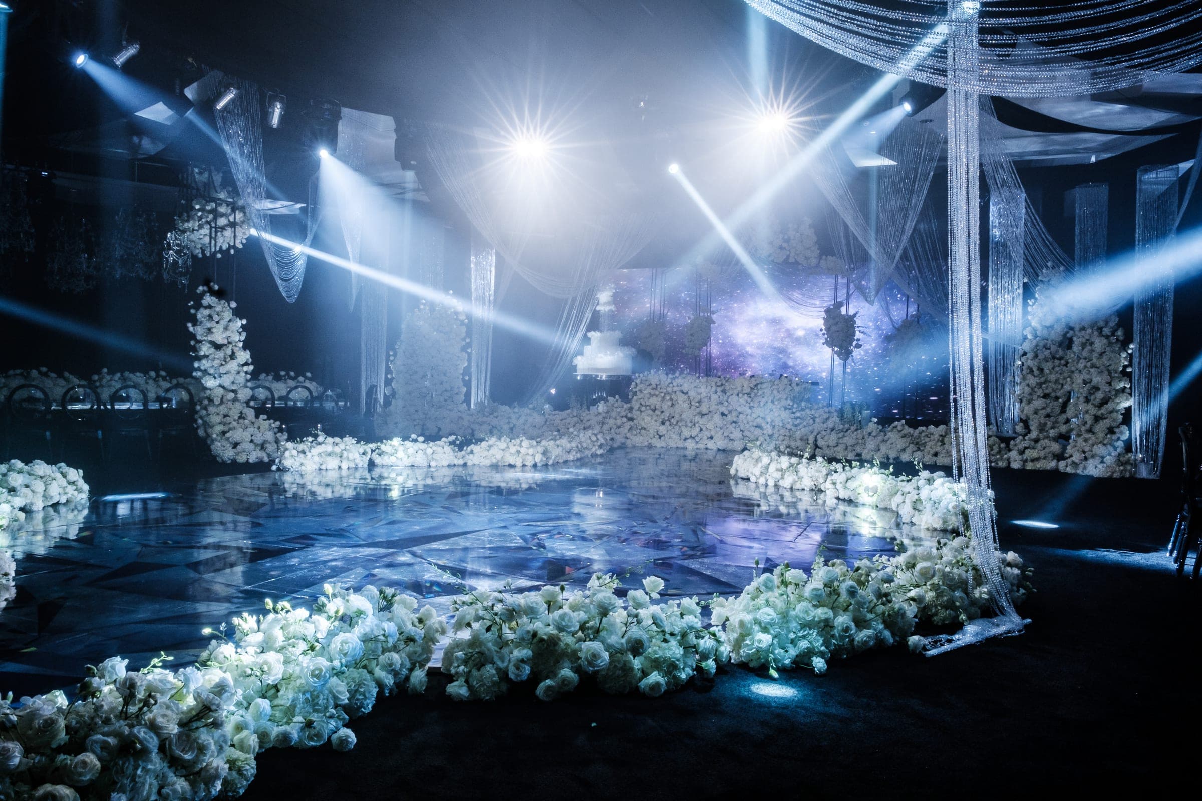 Misty ballroom dance floor edged with white roses under dramatic blue spotlights and beaded drapery.