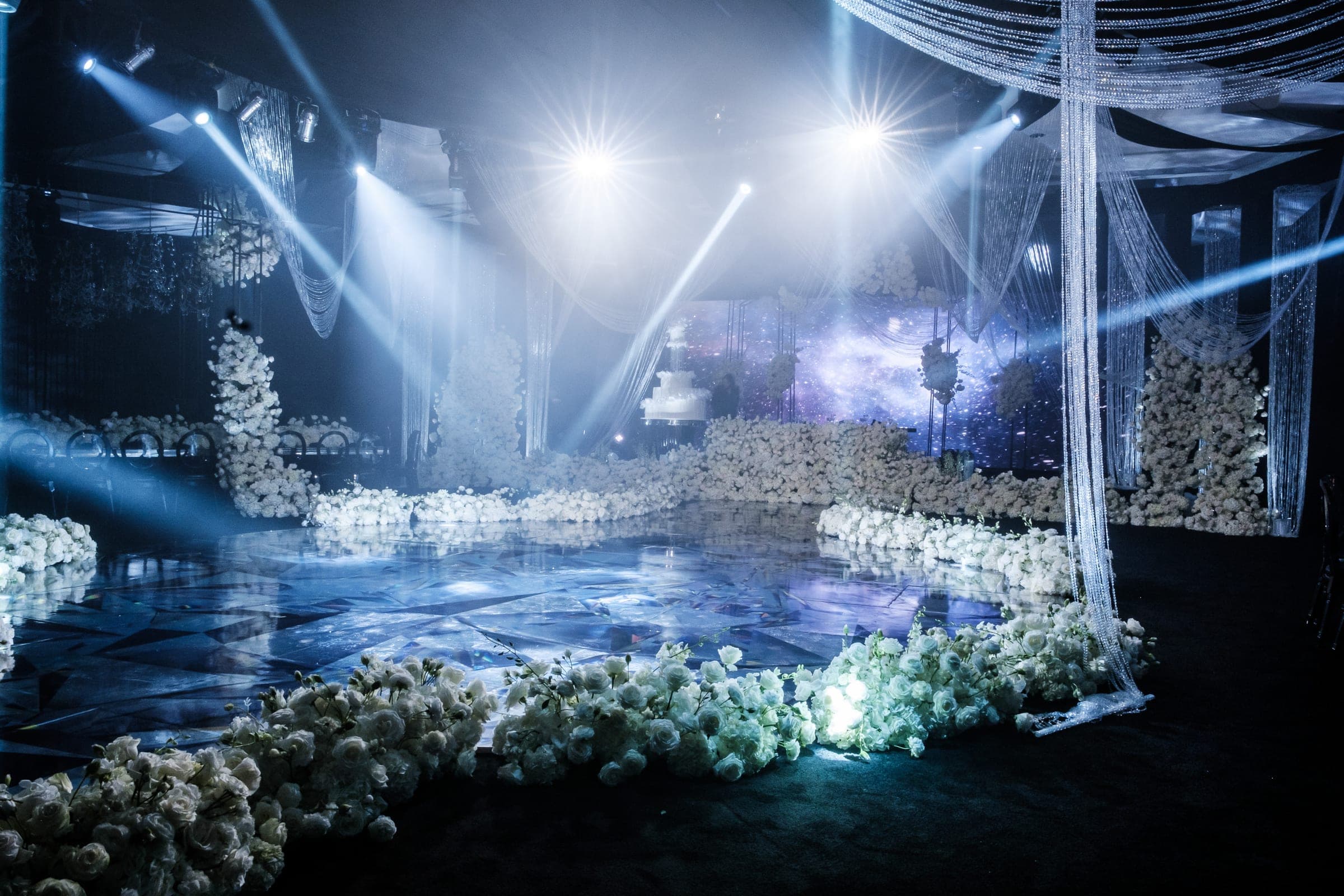 Reflective dance floor surrounded by white floral arrangements glowing under blue and white beams of light.